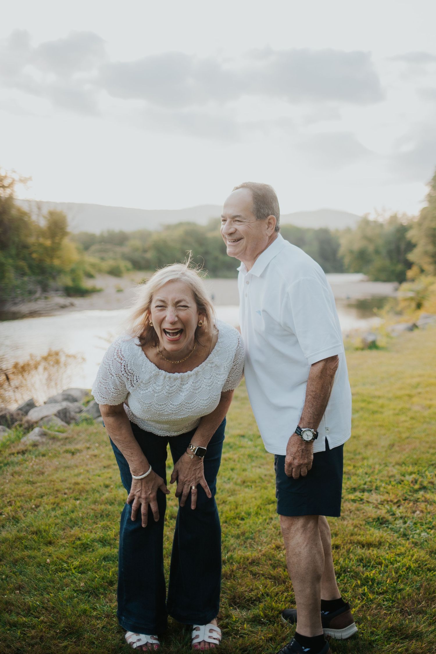 Elderly couple laughing joyfully outdoors by a river with green grass and mountains in the distance.