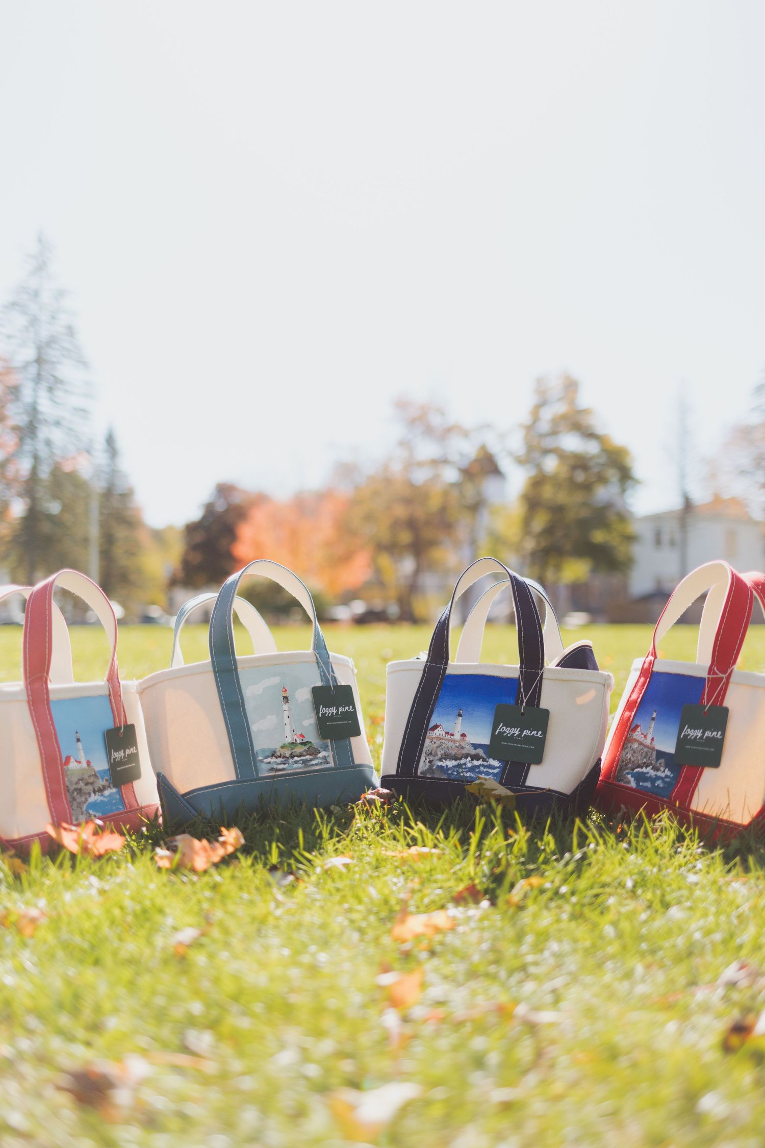 Four canvas tote bags with colorful trim and lighthouse prints sit on a grassy lawn dotted with fallen autumn leaves.