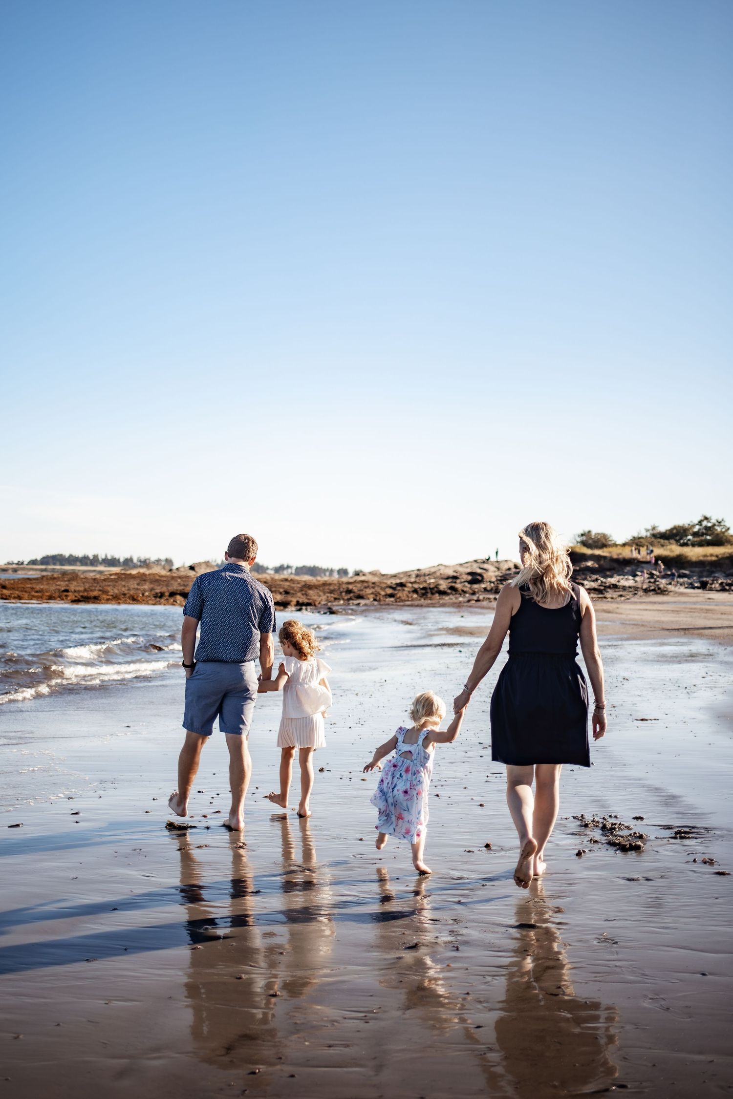 Family of four walking hand-in-hand along a sunny beach shoreline, parents and two young girls enjoying a seaside stroll.