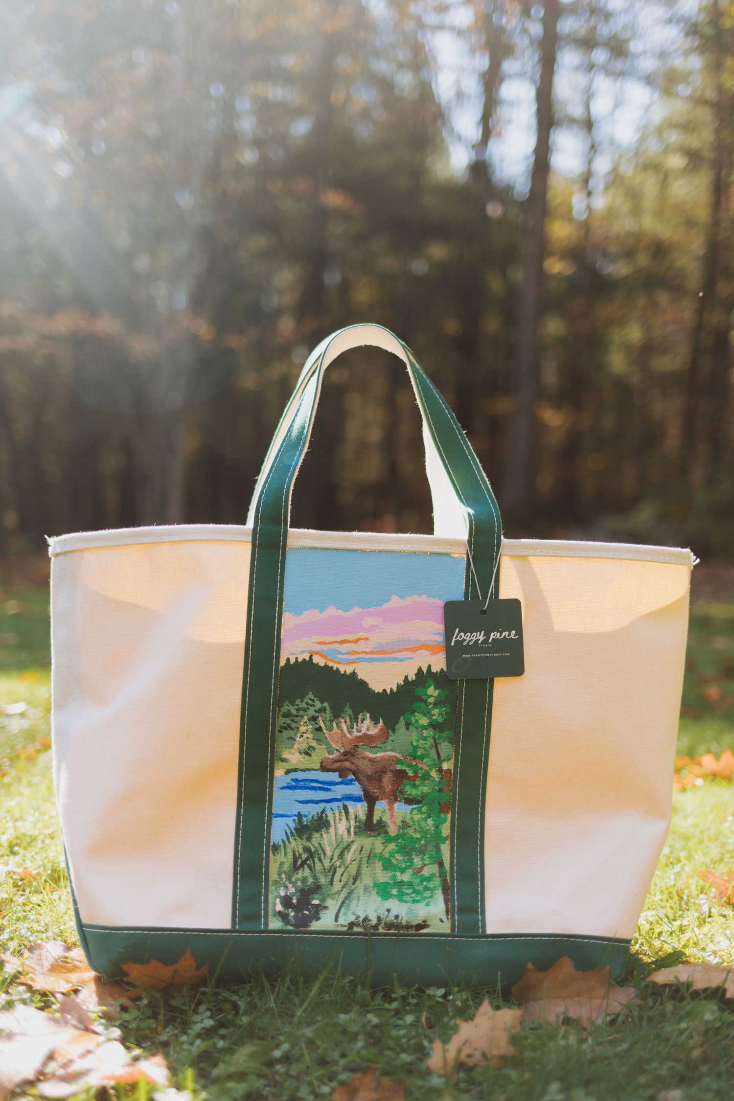 Cream canvas tote bag with green handles and trim, featuring a moose by a lake scene, resting on autumn leaves in a sunlit forest.