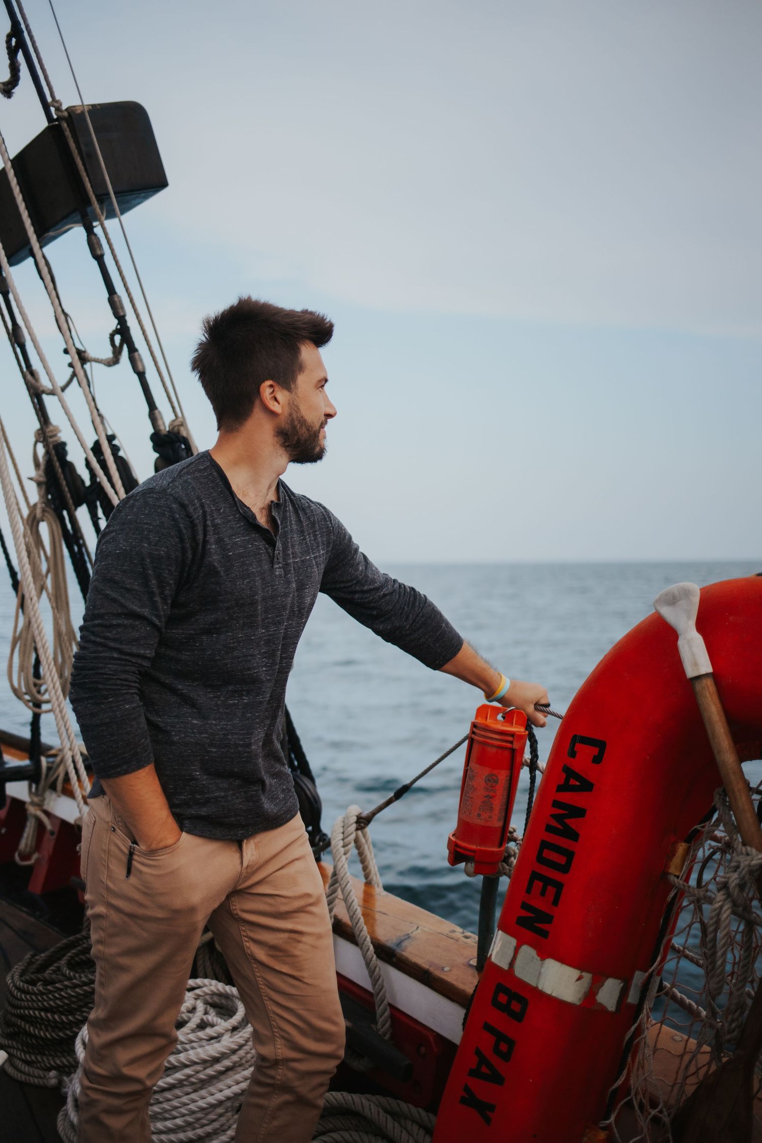 Bearded man in gray shirt standing on a sailboat deck holding a red life preserver ring at sea.