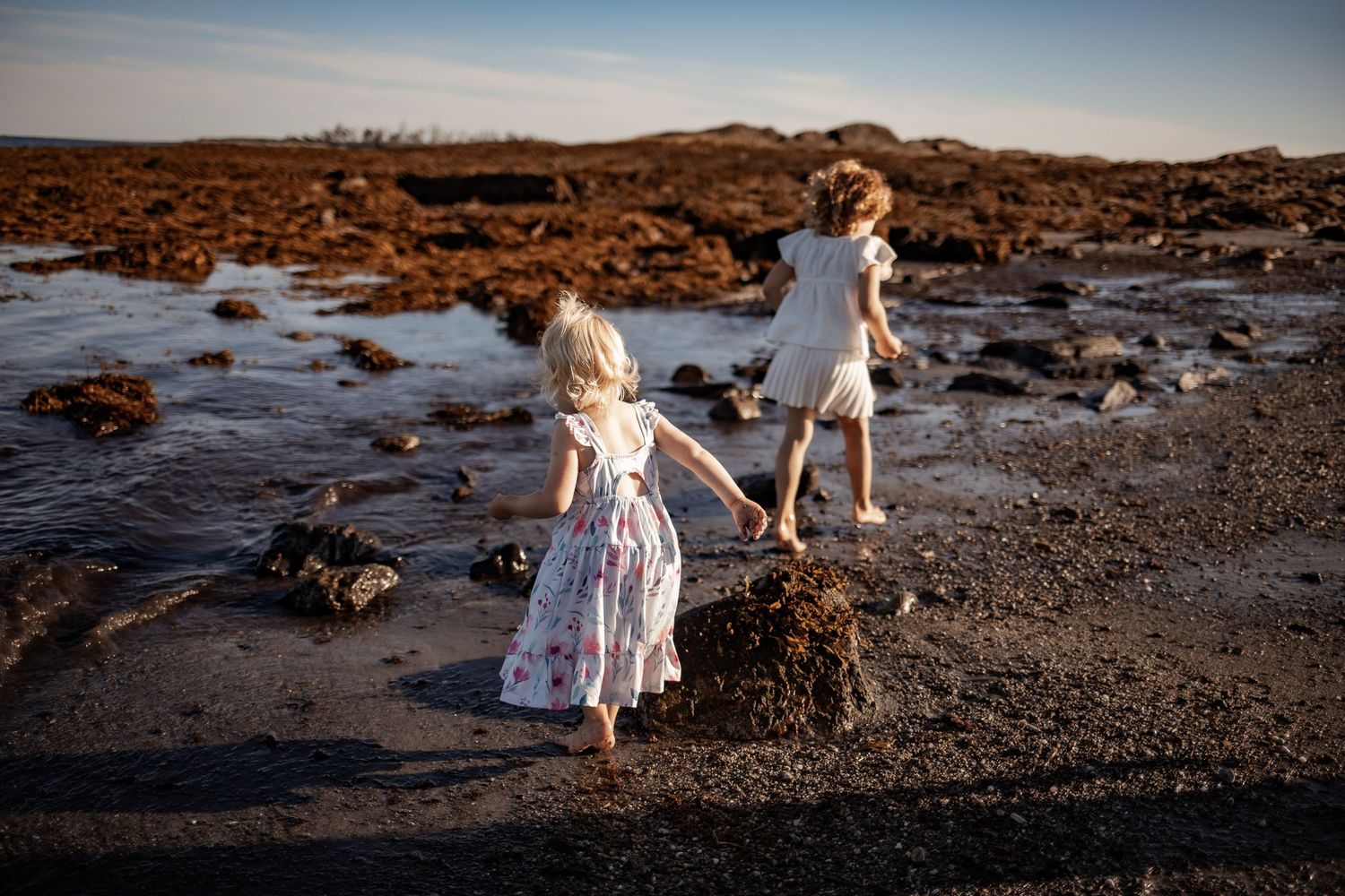 Two young girls in dresses exploring a rocky tidal beach at golden hour, walking through shallow water.