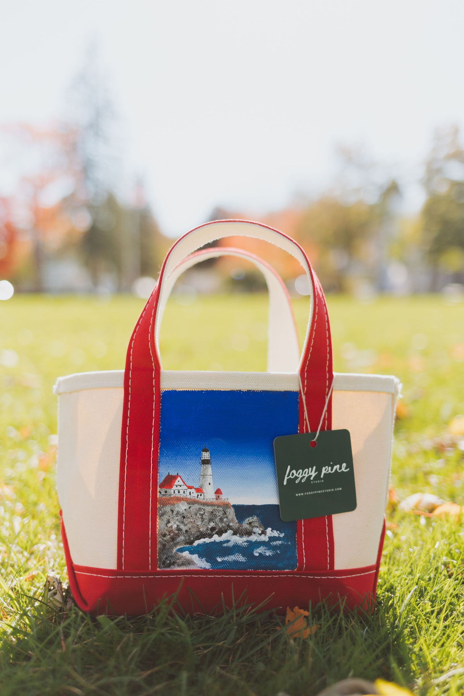 Red and cream canvas tote bag with a hand-painted lighthouse on rocky coast and a Foggy Pine tag, resting on sunlit autumn grass.