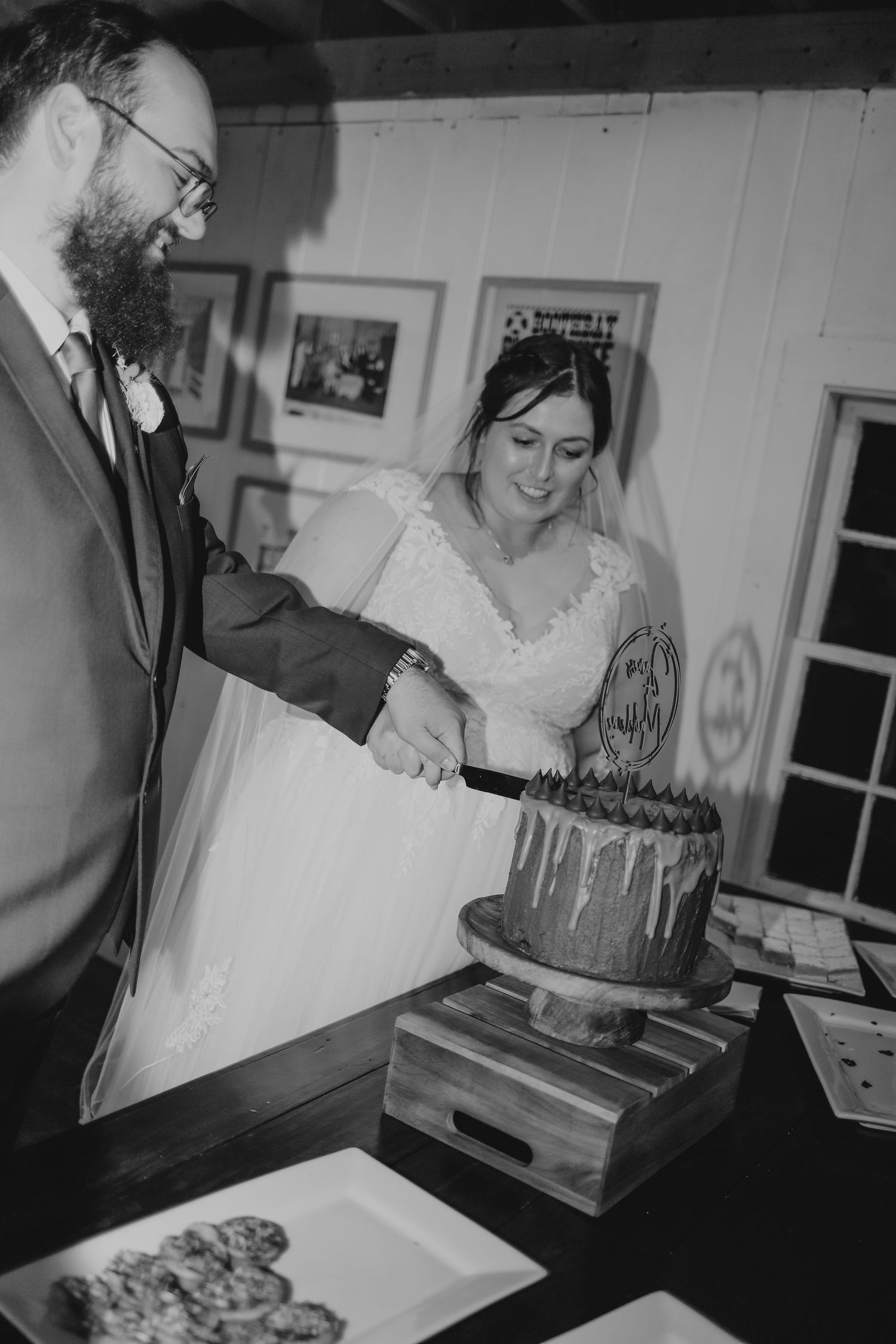 Black and white photo of bearded groom and bride cutting wedding cake at rustic venue.
