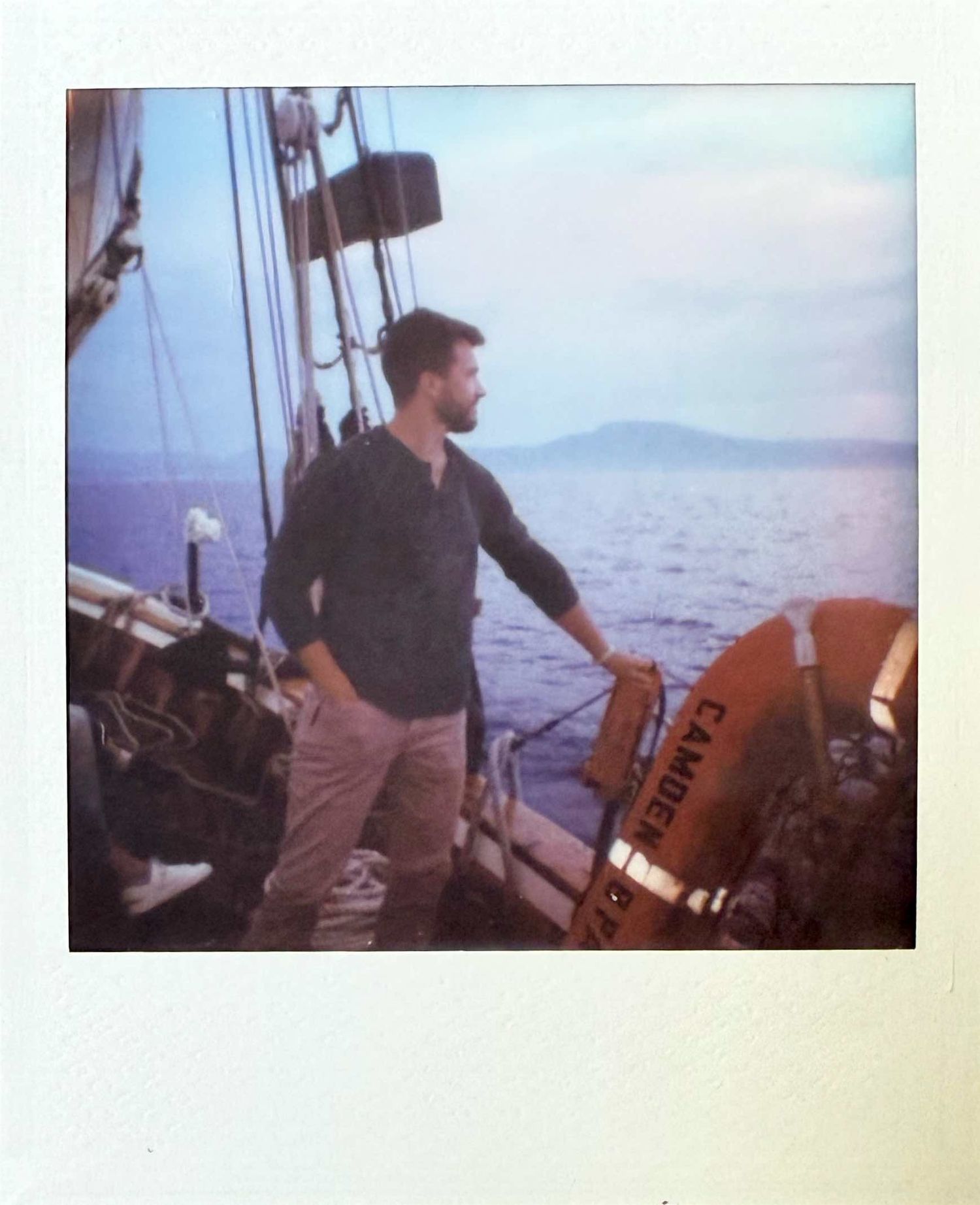 Bearded man standing on a sailboat deck holding a life preserver ring on calm ocean waters.