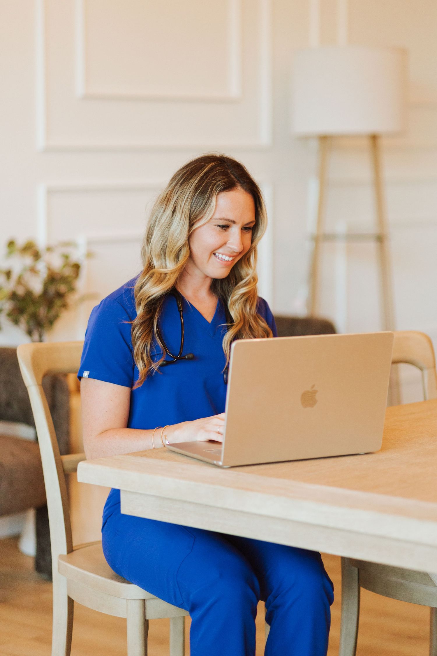 Smiling female nurse in blue scrubs sitting at a wooden table using a gold Apple MacBook laptop.