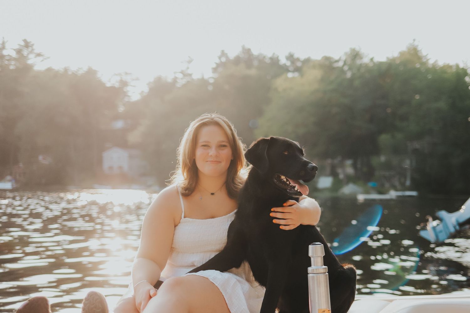 A smiling woman sits on a dock with a black Labrador dog by a sunlit lake.