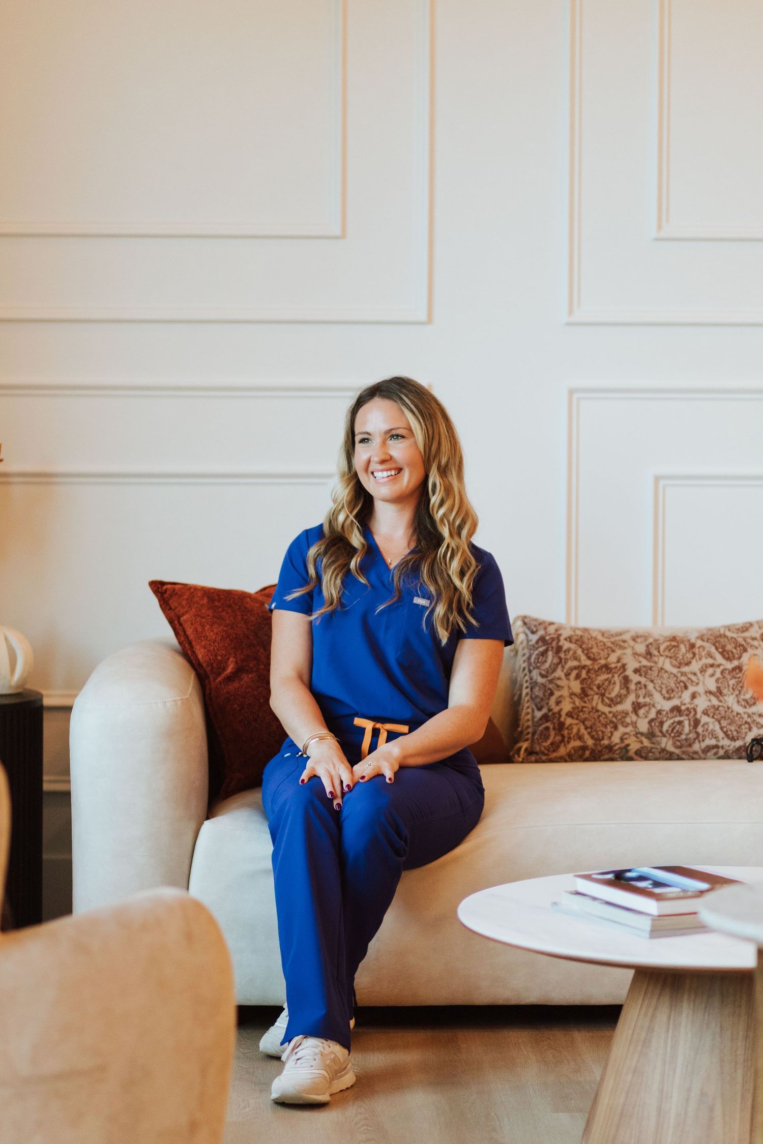 Smiling woman with long wavy hair in blue scrubs sits on a cream sofa with patterned pillows in a bright, modern room.