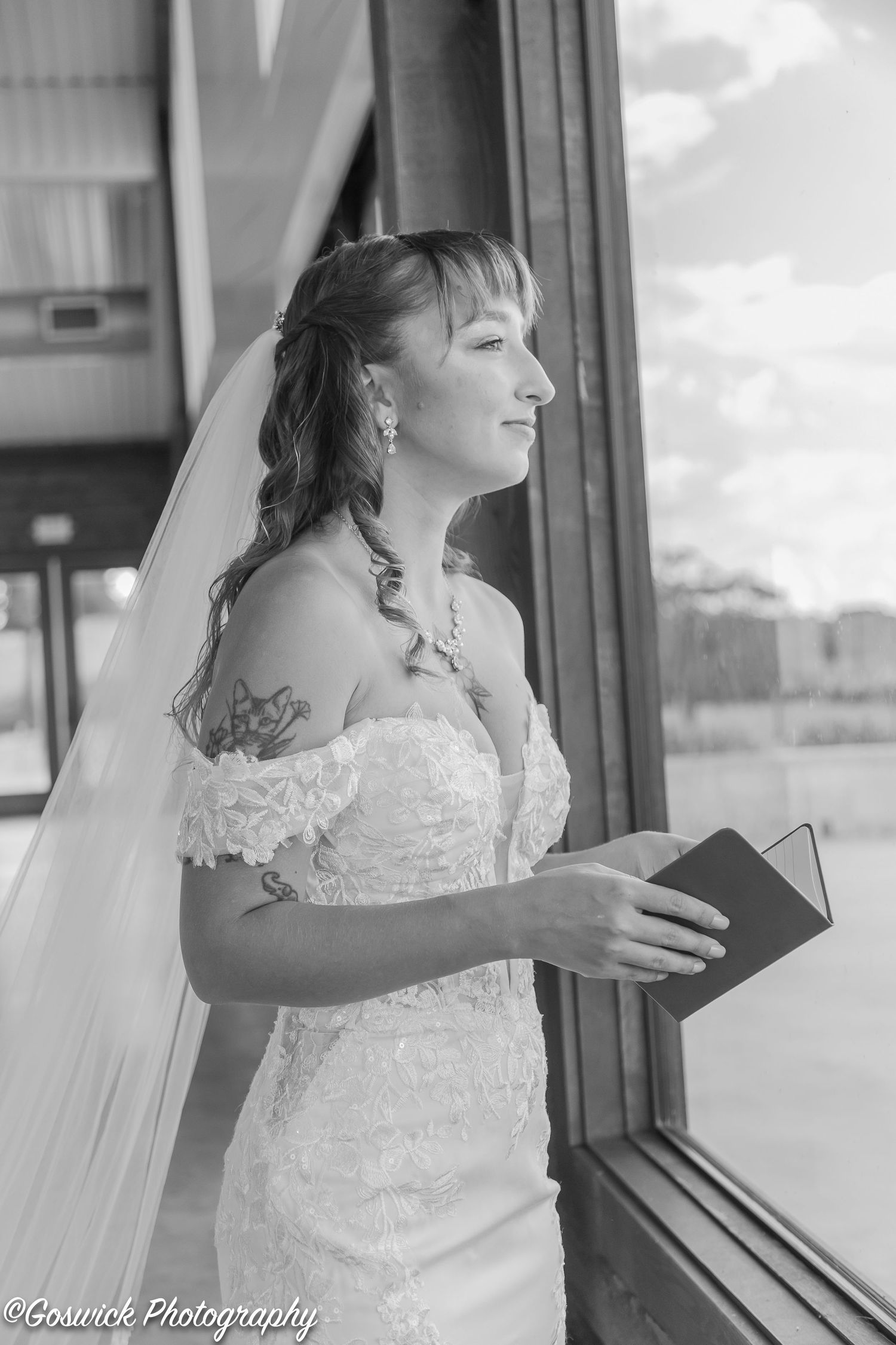 Black and white portrait of bride with tattoo holding vow book while gazing out a window.