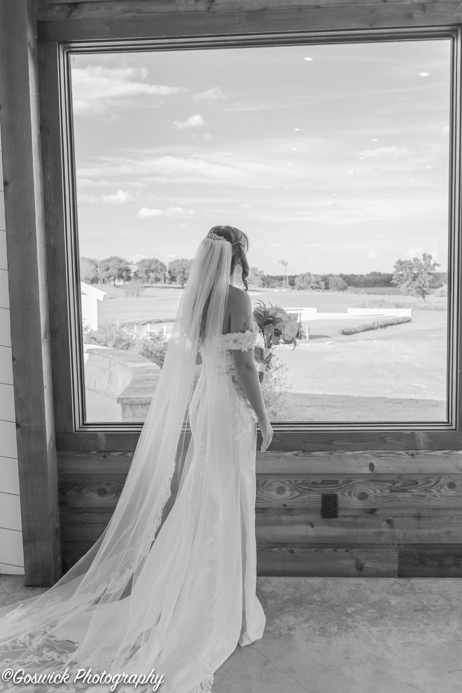 Black and white photo of bride in lace gown with veil gazing out a large window at countryside.