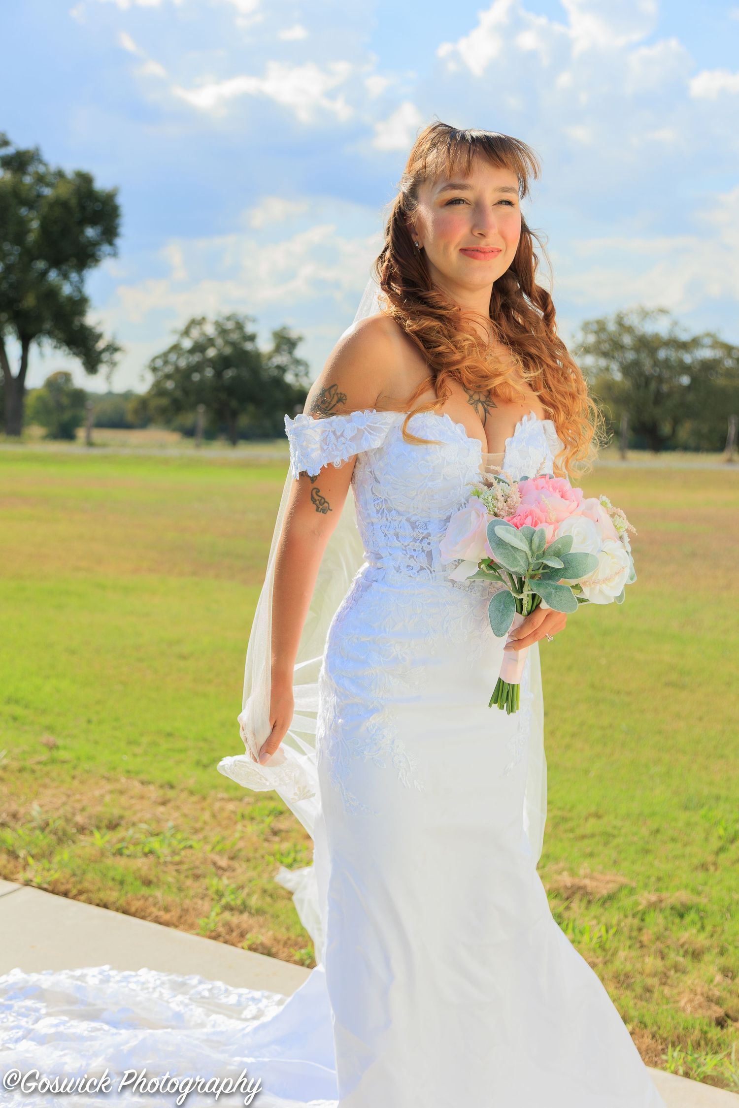 Smiling bride in off-shoulder lace mermaid gown holds pink bouquet outdoors on sunny day with green field.