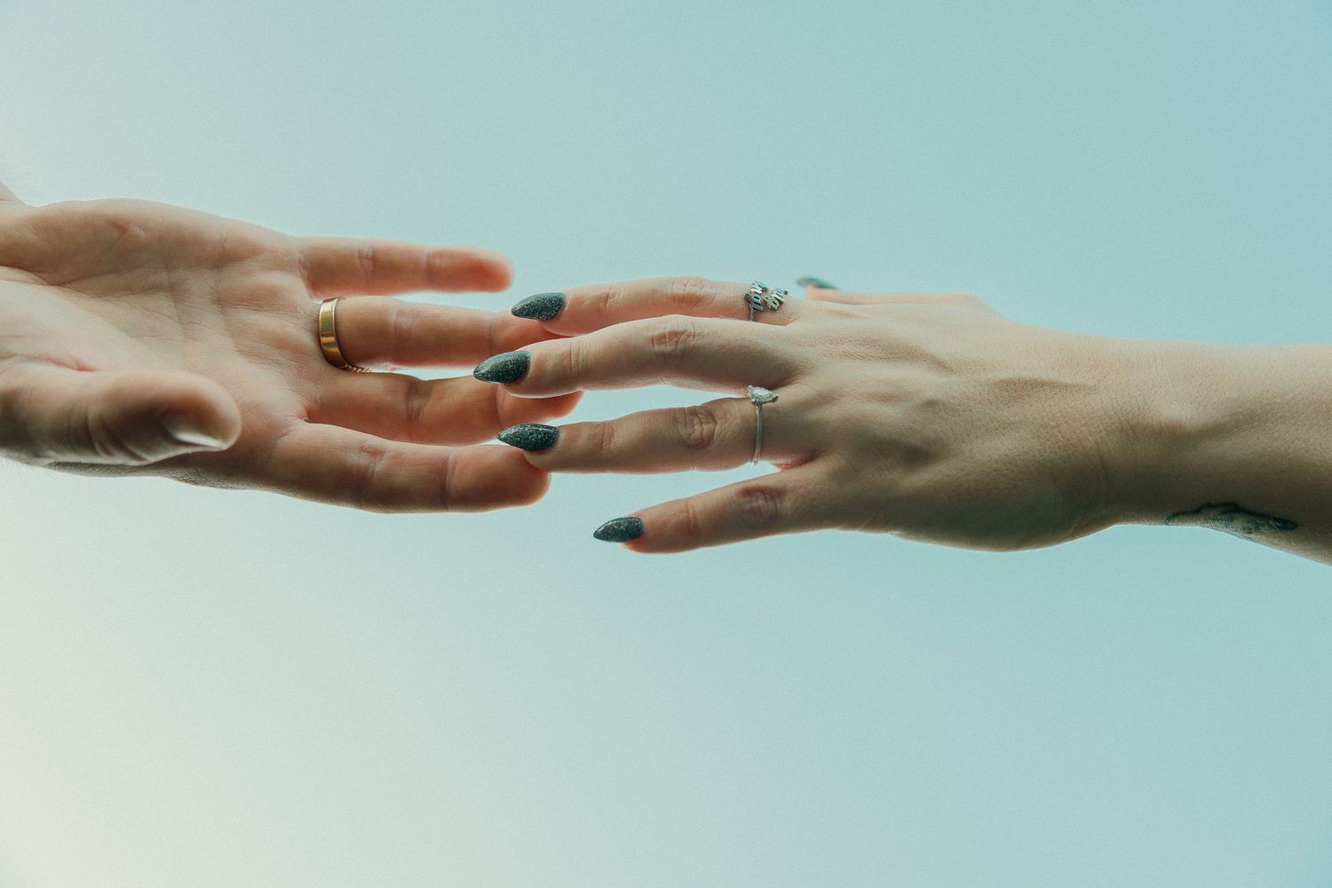 Two hands with rings and dark nail polish reaching toward each other on blue background.
