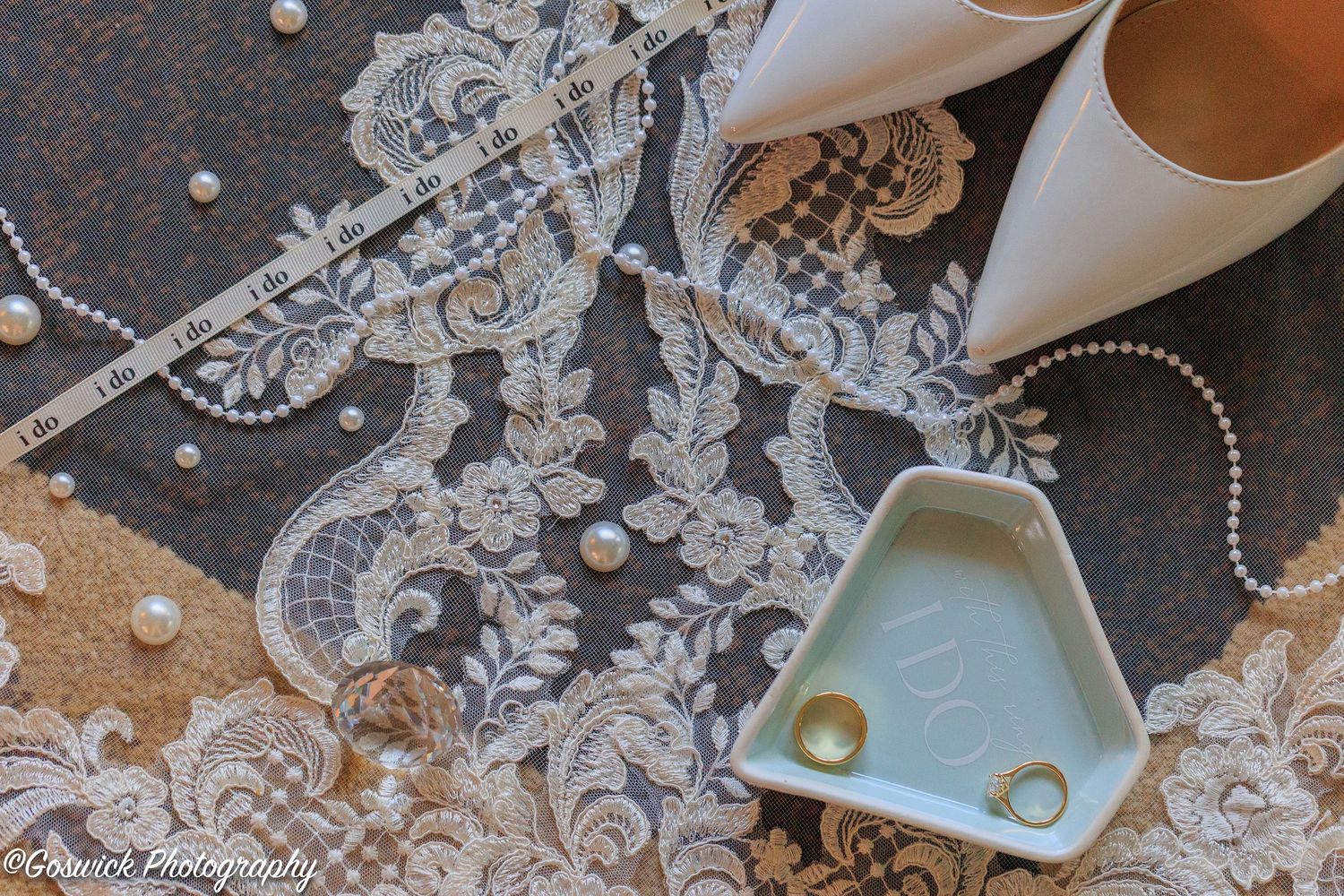 Bridal detail flat lay with lace veil, pearl necklace, gold rings in a ceramic dish, and white heels.