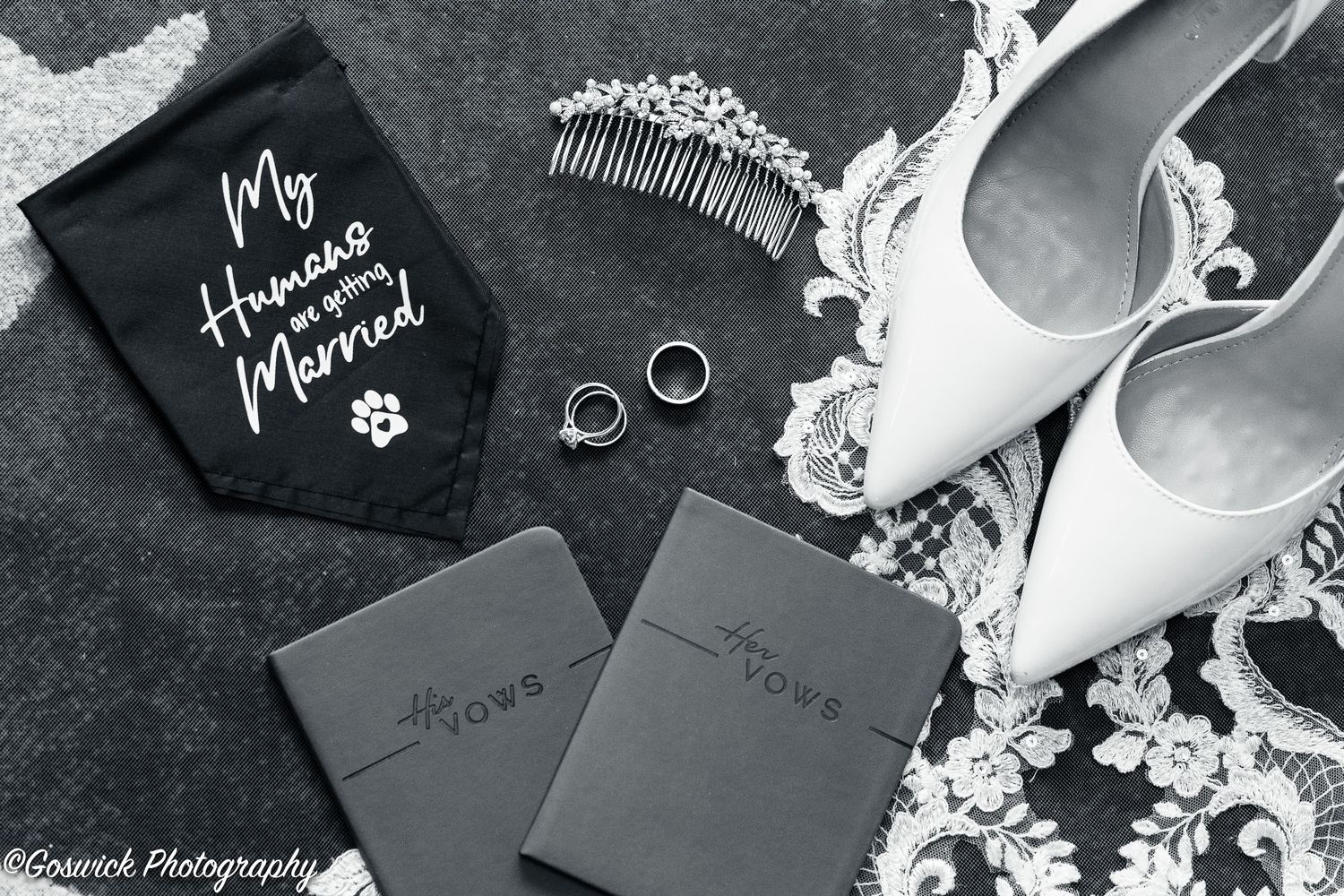 Black and white flat lay of bridal accessories including heels, rings, and vow books.