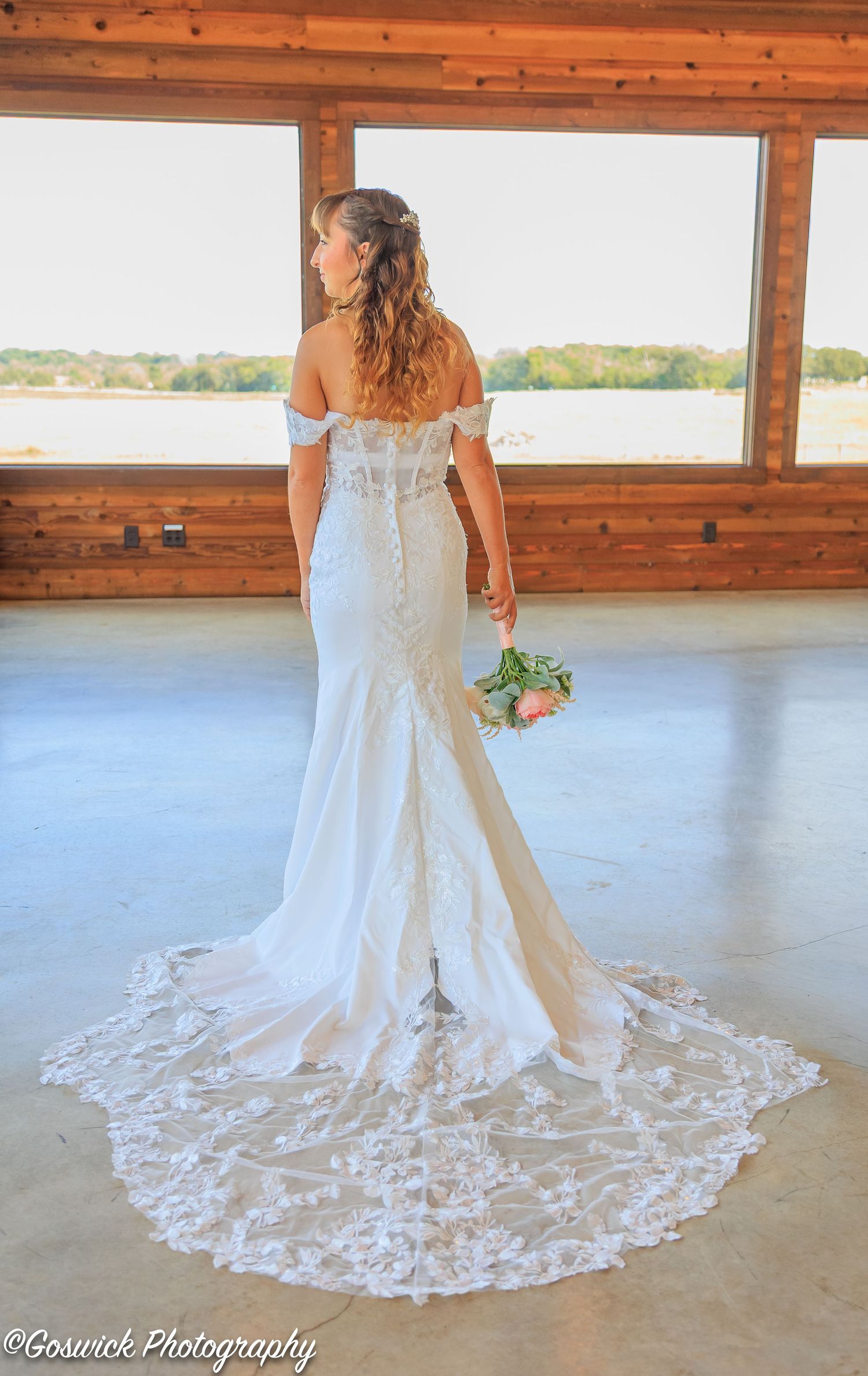 Bride in off-shoulder lace mermaid wedding gown with long train stands facing window in rustic barn venue.