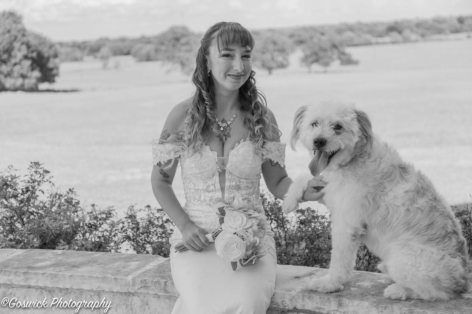 Black and white portrait of smiling bride in lace gown posing with fluffy white dog in scenic outdoor setting.