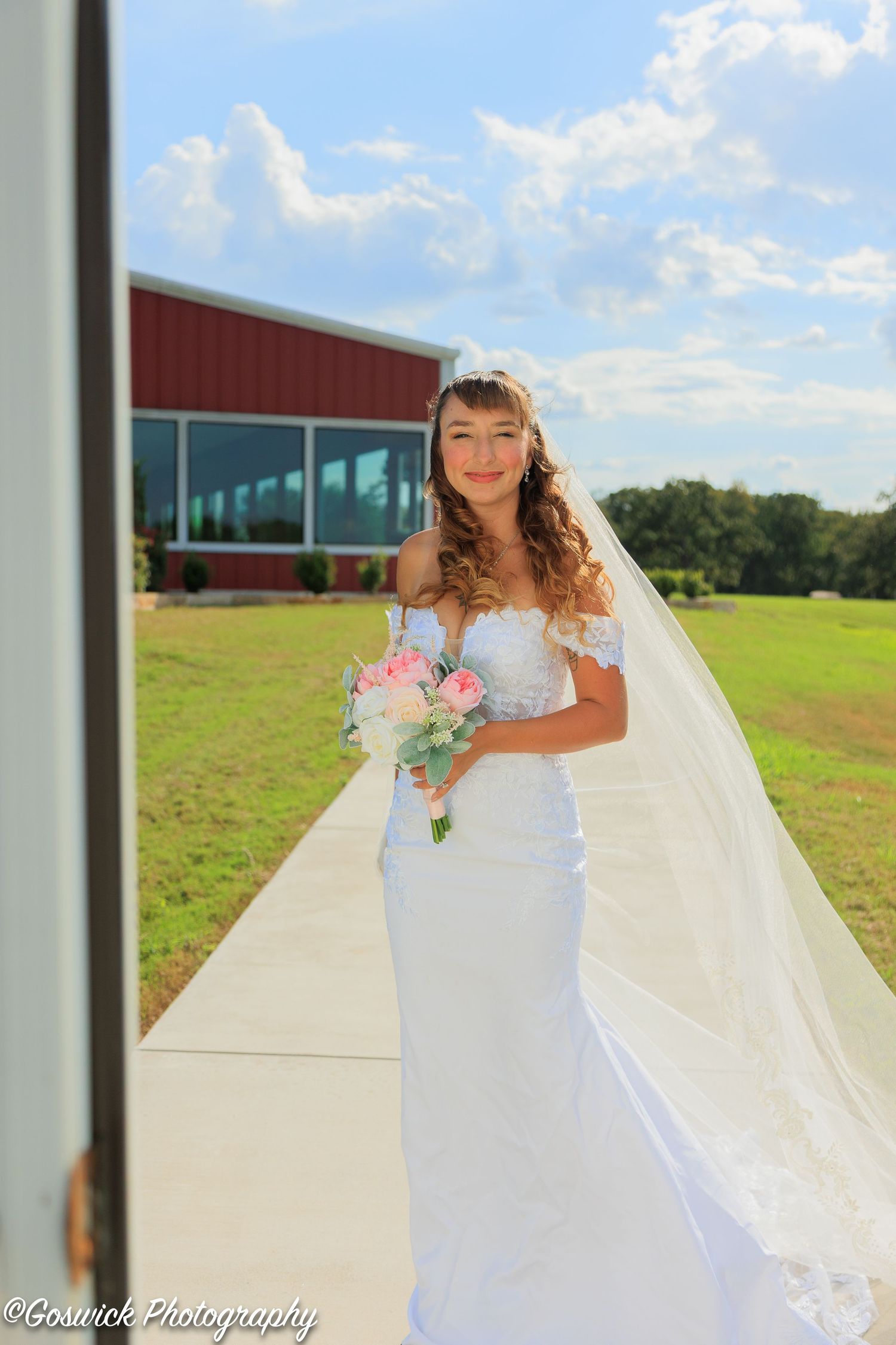 Bride in off-shoulder lace wedding gown with veil holds pink bouquet on sidewalk outside red barn venue.
