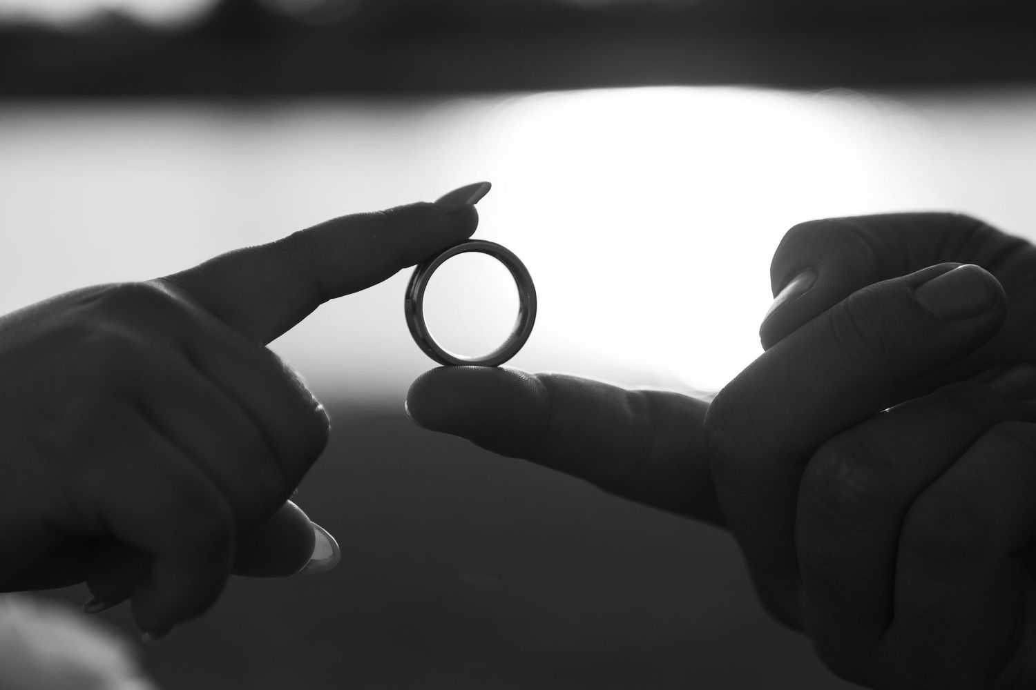 Black and white photo of two hands holding a wedding ring between their fingertips against a softly blurred sunset background.