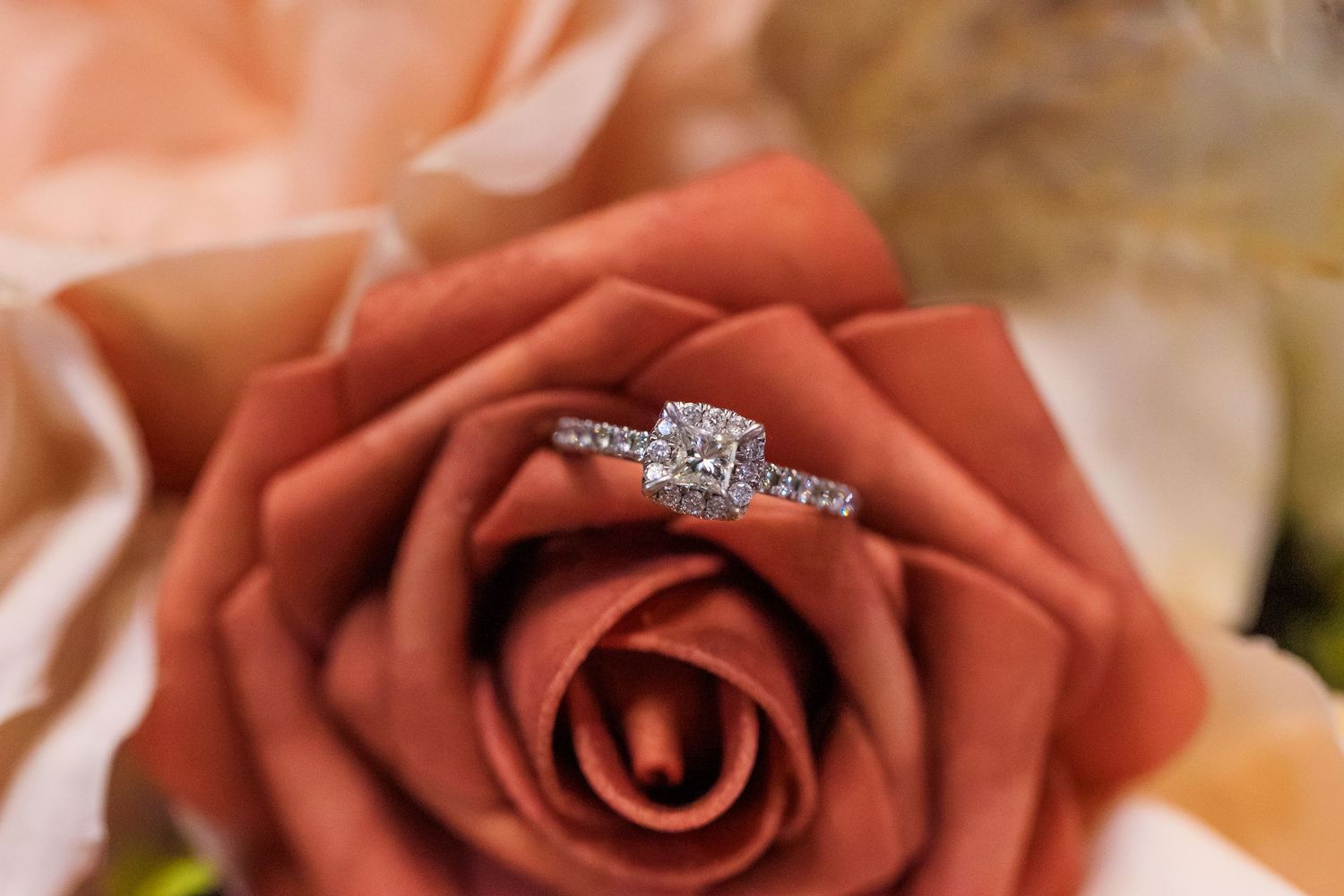 Diamond engagement ring with a square center stone and pavé band resting on a terracotta rose surrounded by soft peach flowers.