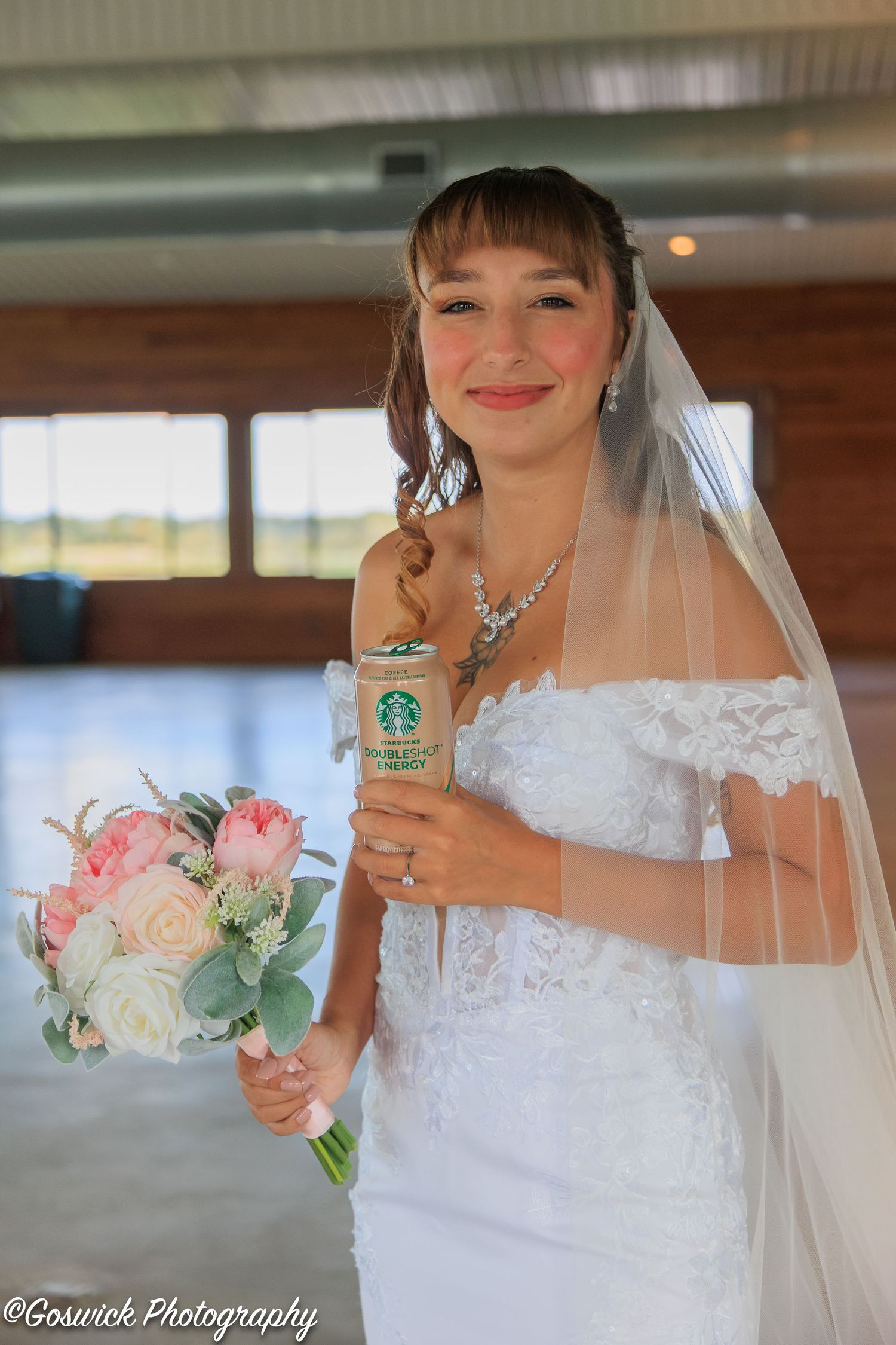Smiling bride in off-shoulder lace gown holding a bouquet and Starbucks drink on her wedding day.