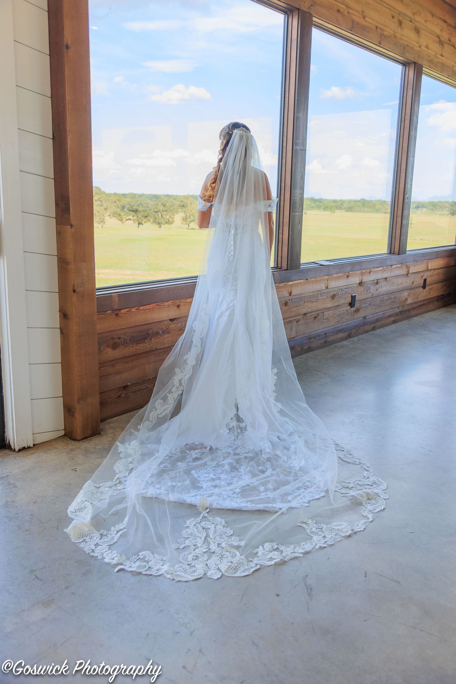 Bride in off-shoulder lace gown with stunning cathedral veil overlooking a scenic countryside view.
