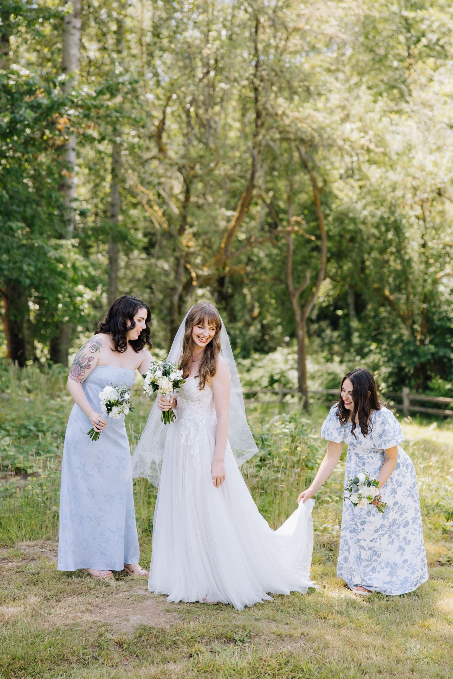 Bride in white gown and veil stands with two bridesmaids in blue and floral dresses in a lush garden in Bay Area.