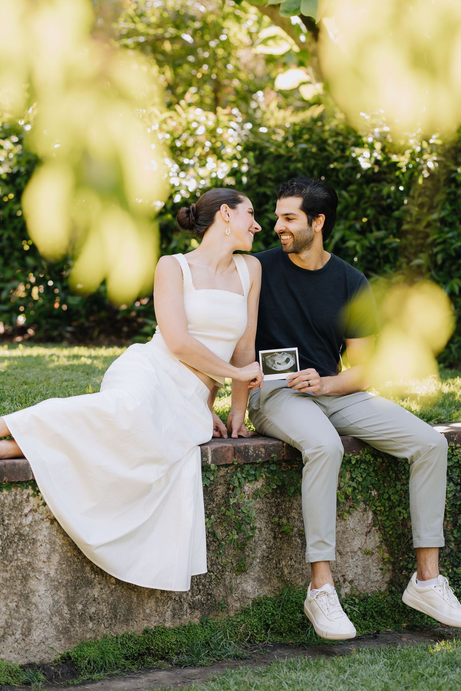 Smiling couple sitting on a stone ledge in a sunny garden, holding an ultrasound photo together at Filoli Woodside maternity photoshoot.