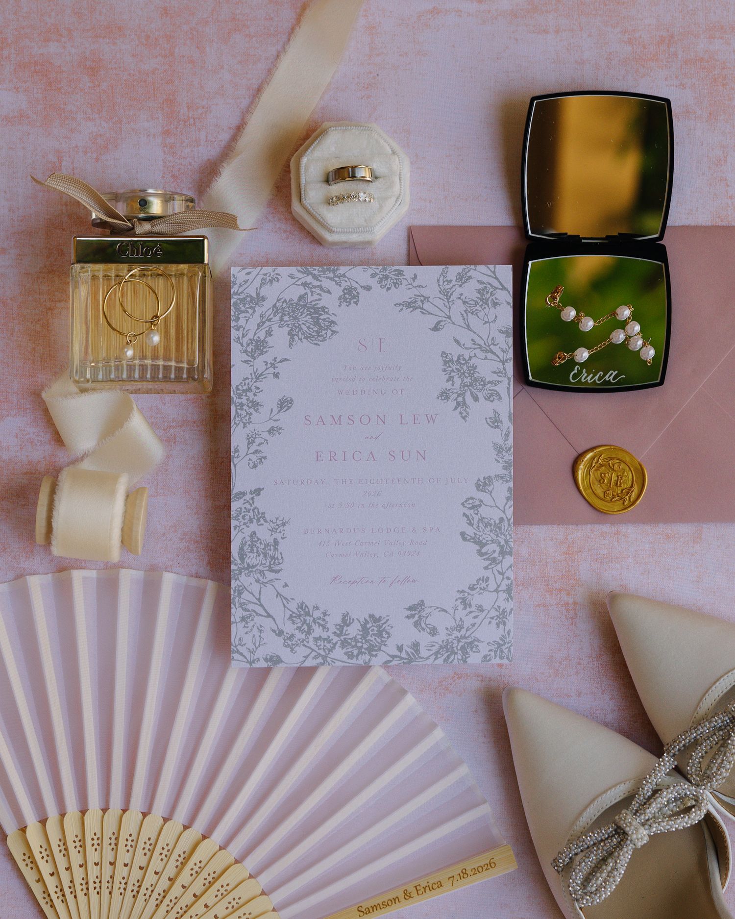 Elegant wedding stationery flatlay with floral invitation, perfume, ring box, and blush pink accents.