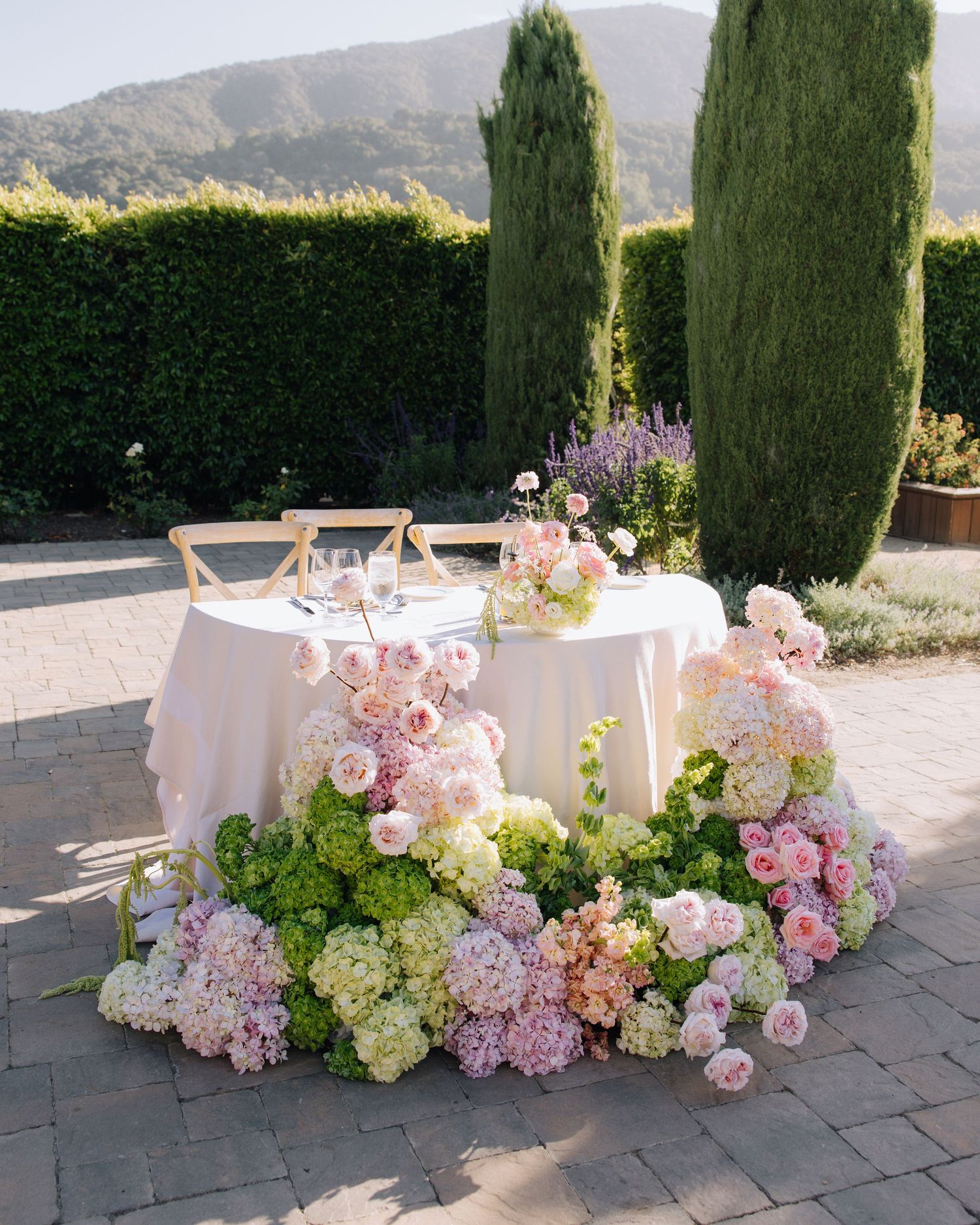 Romantic sweetheart table surrounded by lush pink hydrangea and rose floral ground arrangement outdoors.