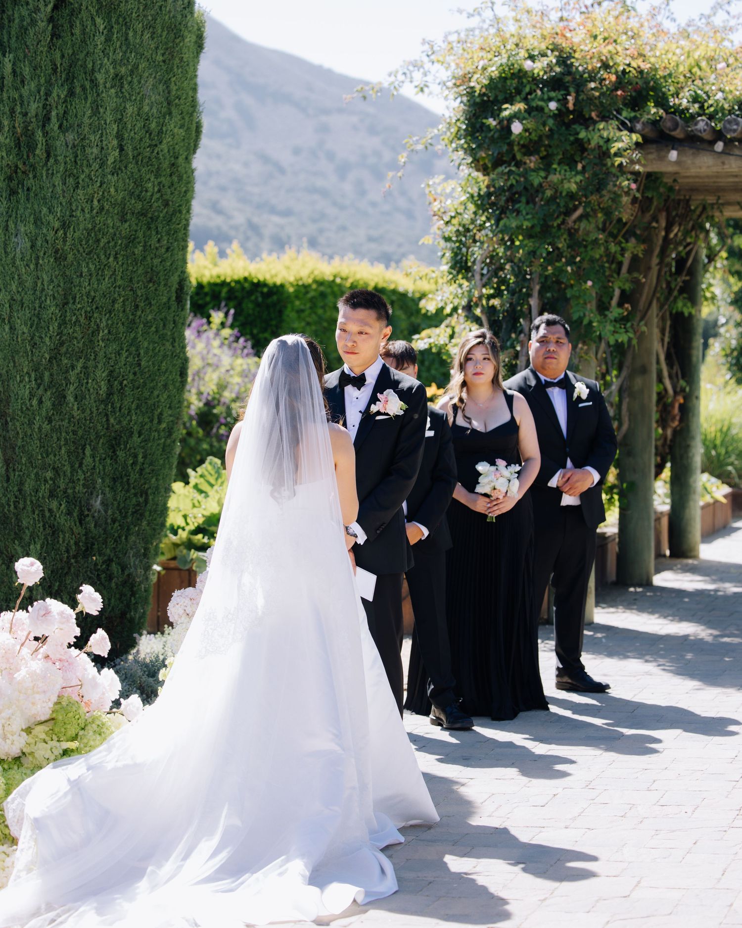 Bride approaching groom at outdoor garden wedding ceremony with cypress trees and mountains at Bernardus Lodge in Carmel Valley, CA.