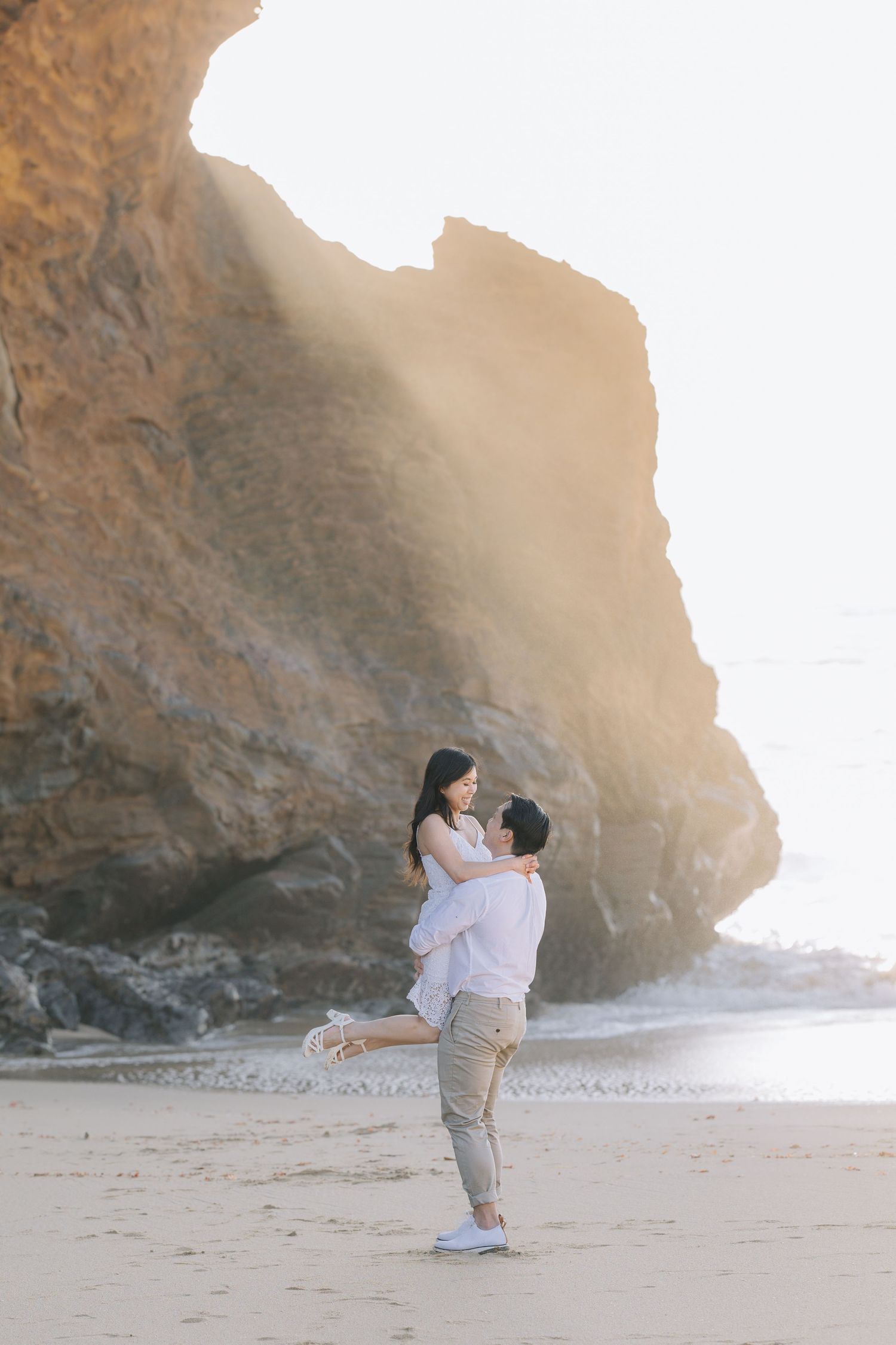 Man lifting woman in white dress on sandy beach with dramatic rock formation behind at Santa Cruz engagement photoshoot.