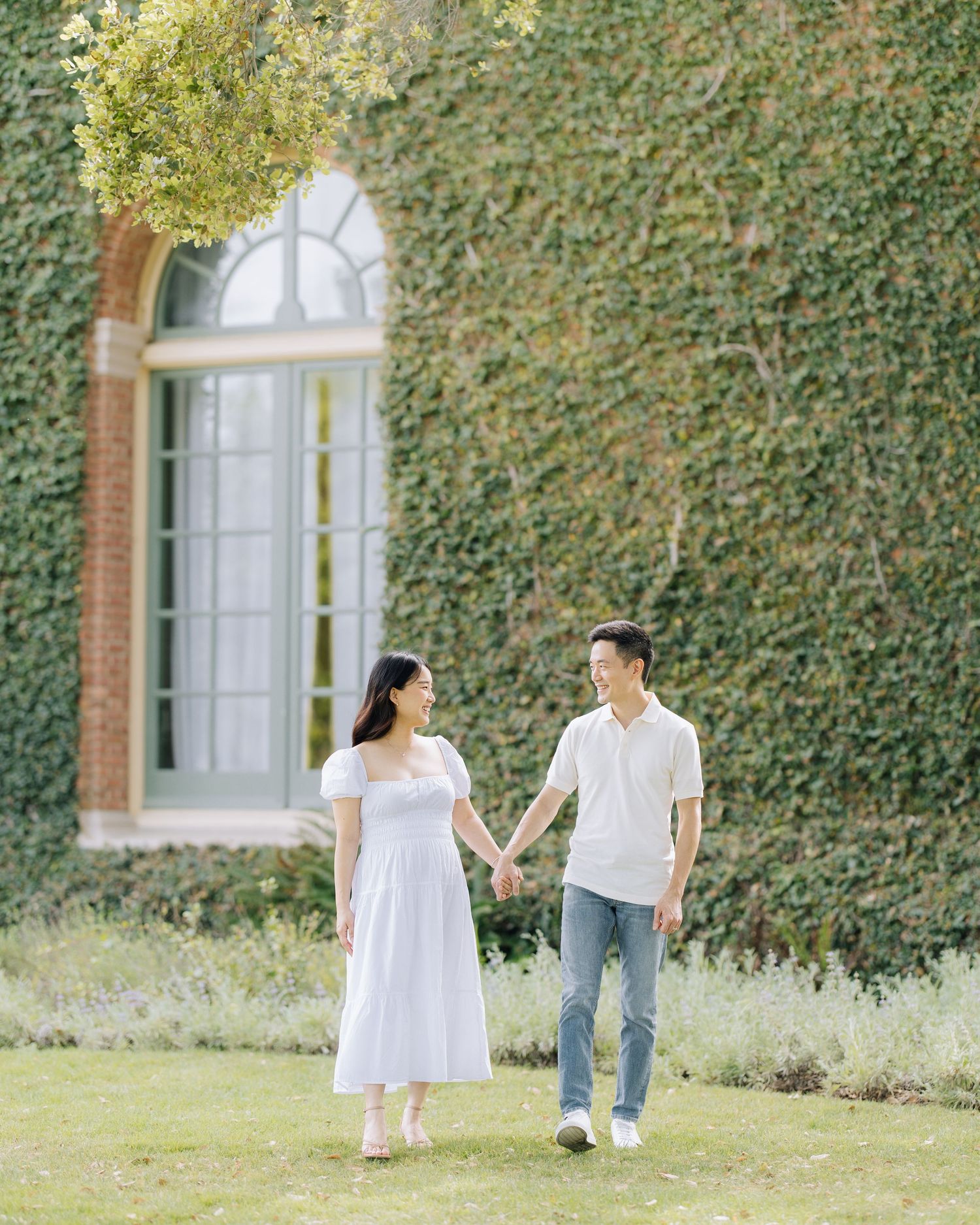 Pregnant couple holding hands walking in garden with ivy-covered brick wall backdrop at Filoli Woodside maternity photoshoot.