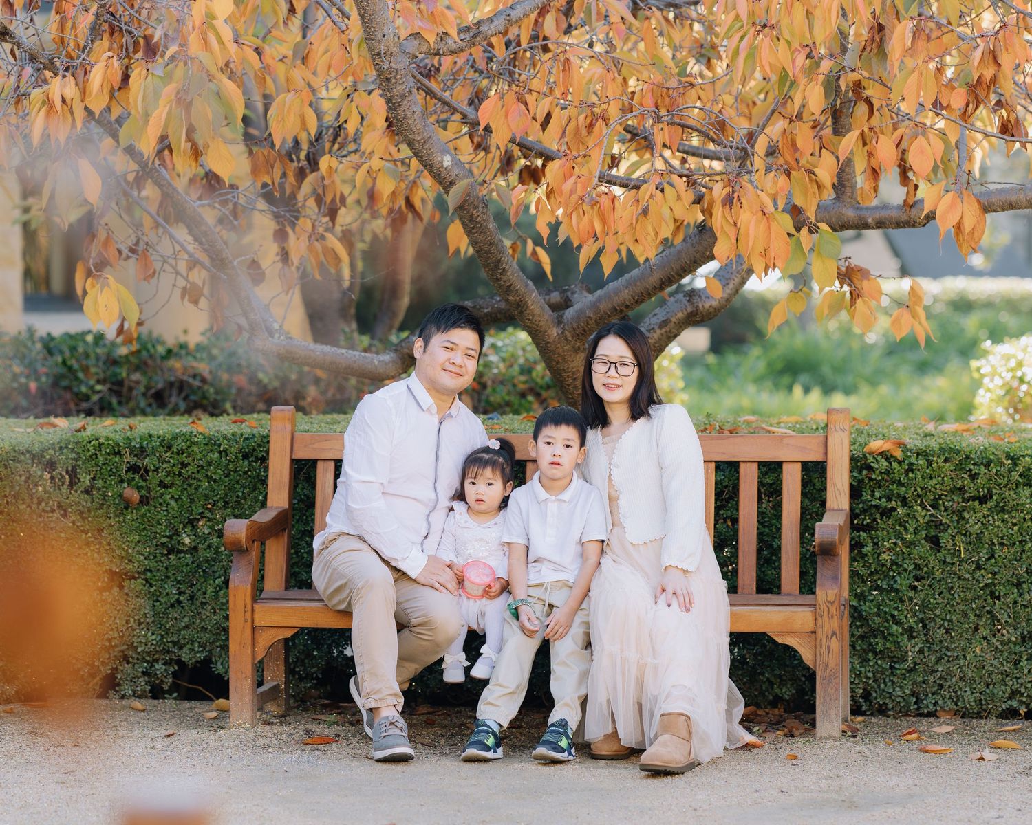 Asian family of four posing on wooden bench under autumn foliage in a garden during Stanford family photoshoot.
