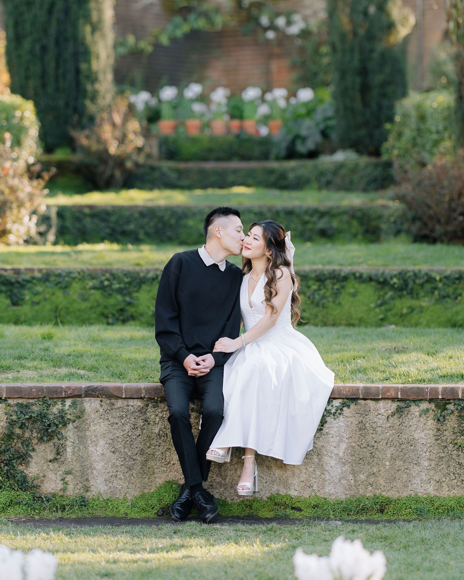 Couple sharing a kiss while seated on a stone wall in a lush formal garden with tulips during Filoli engagement photoshoot.