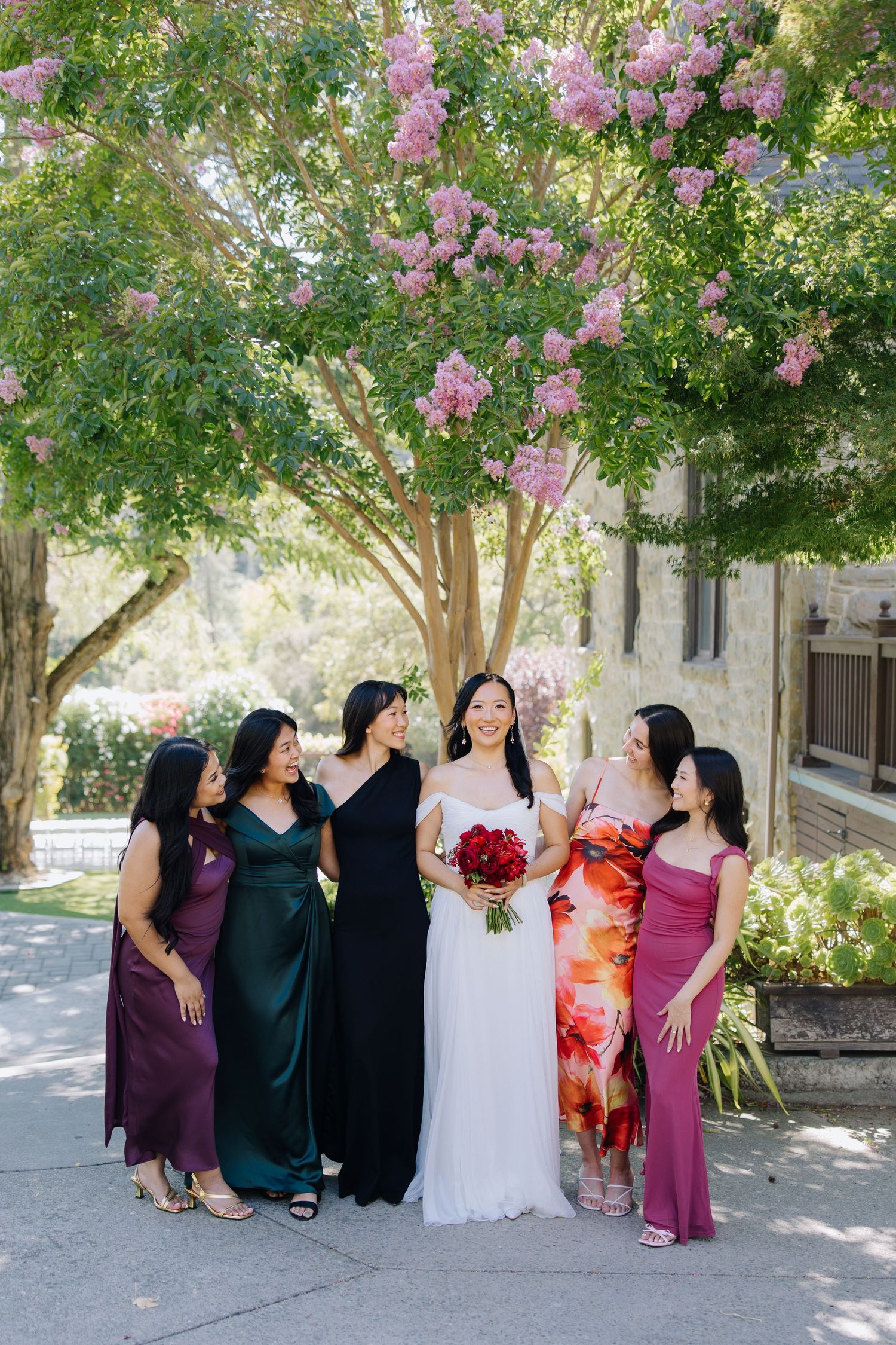 Bride in white gown holds red bouquet smiling with five bridesmaids in colorful dresses under blooming tree at Elliston Vineyards in Sunol,