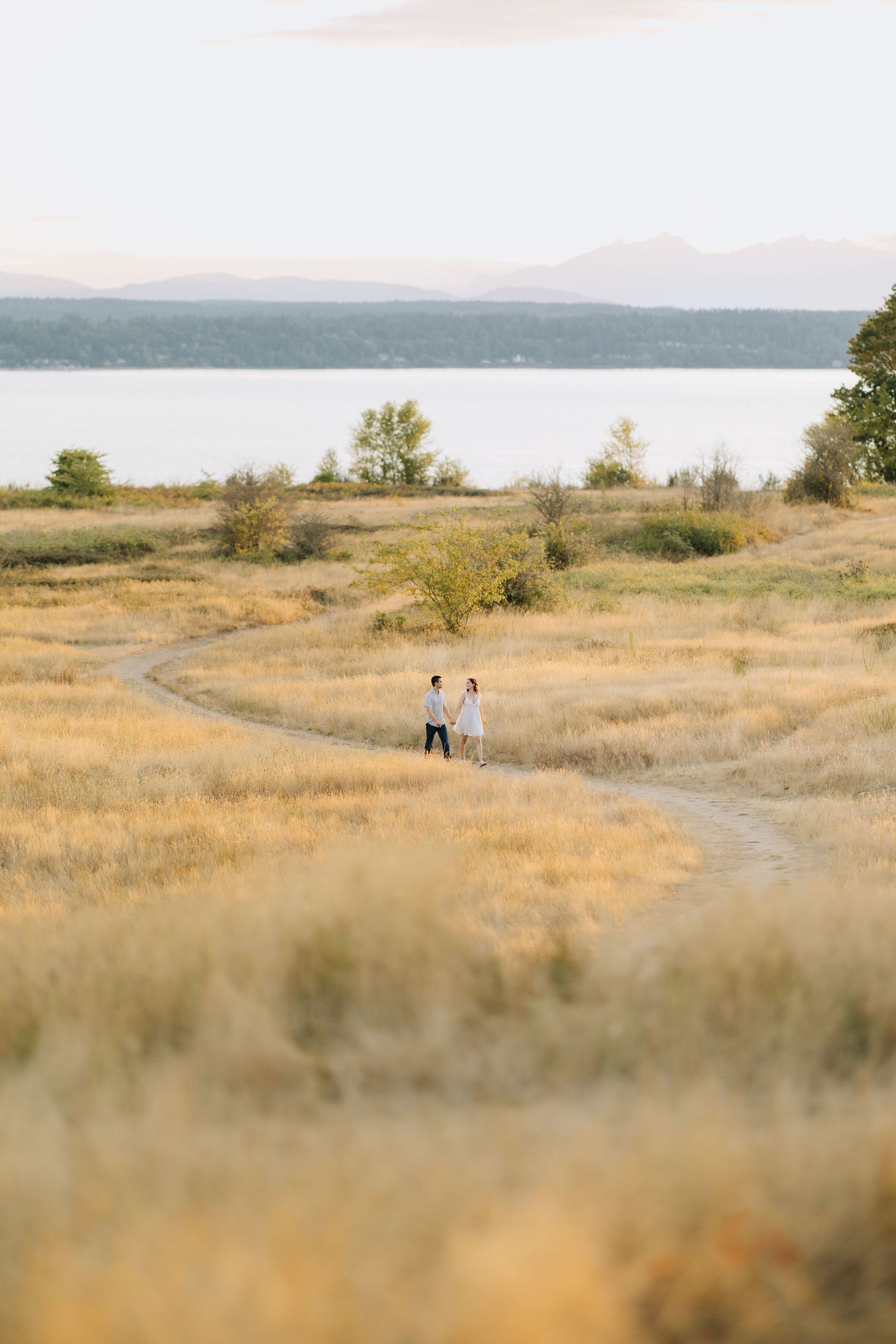 Couple walking hand in hand on winding path through golden meadow with lake and mountains. Engagement session at Discovery Park Seattle.