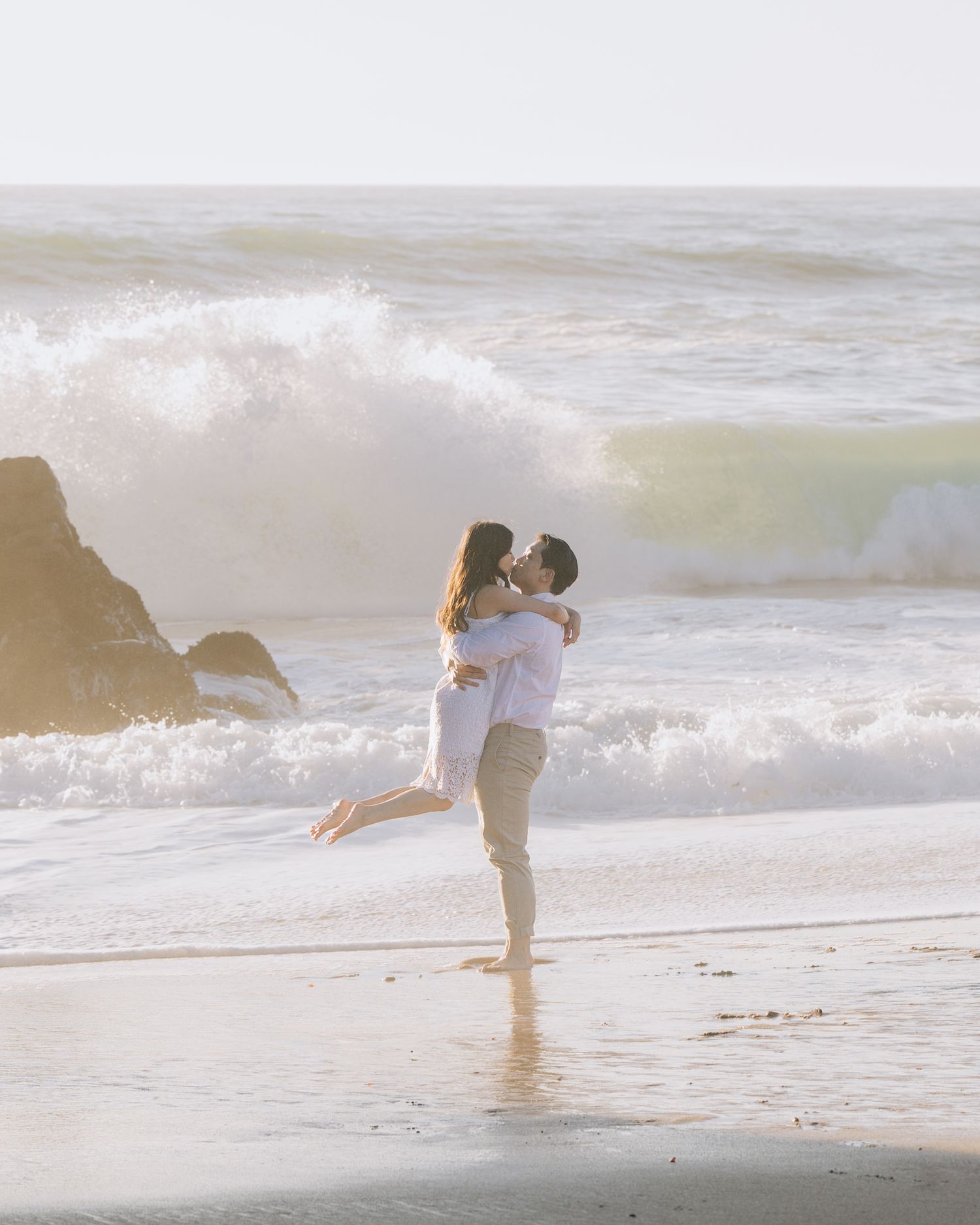 Couple kissing while man lifts woman on beach with crashing waves in background at Santa Cruz engagement photoshoot.