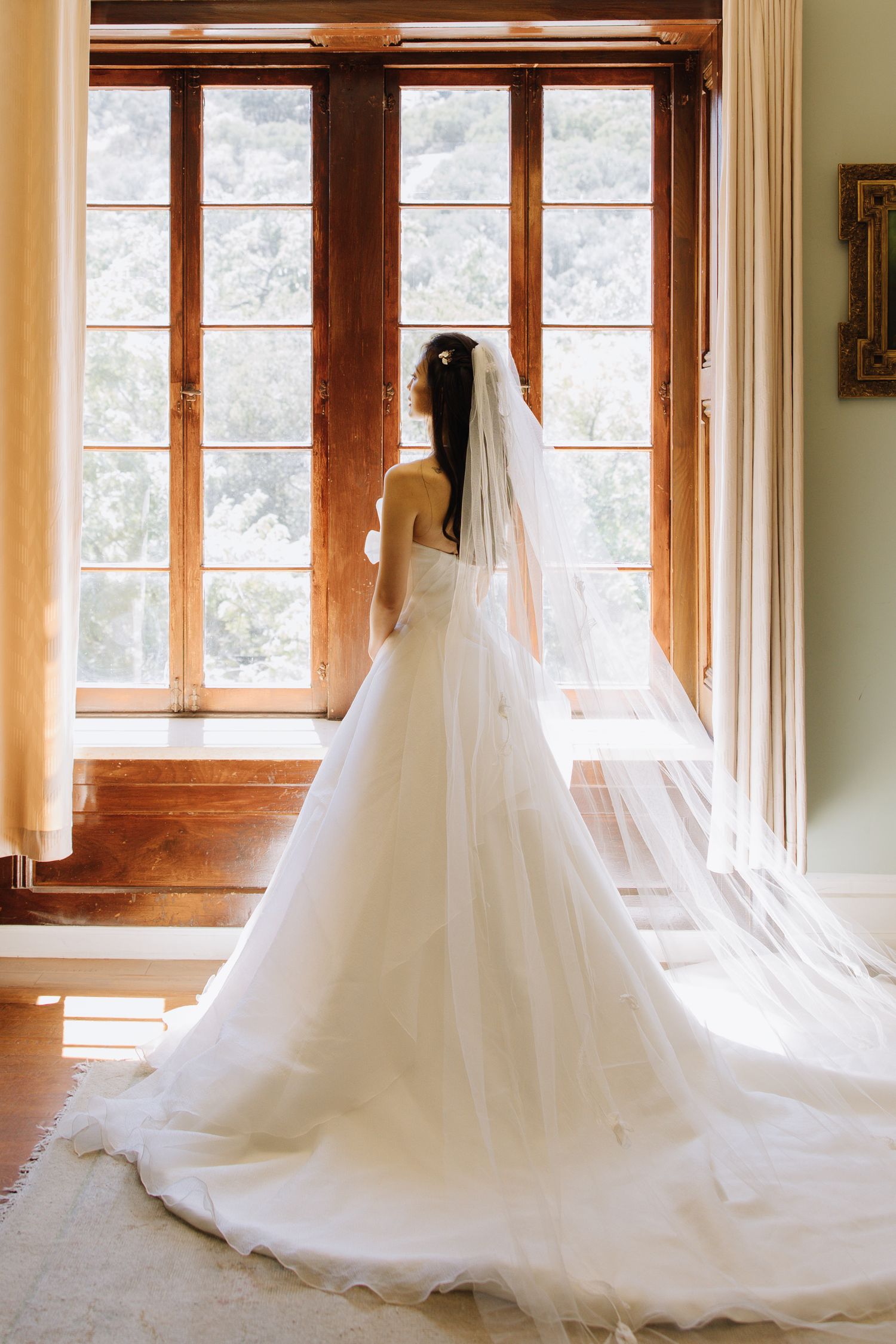 Bride in elegant white ballgown with long cathedral veil stands before large wooden windows at Elliston Vineyards in Sunol, California.