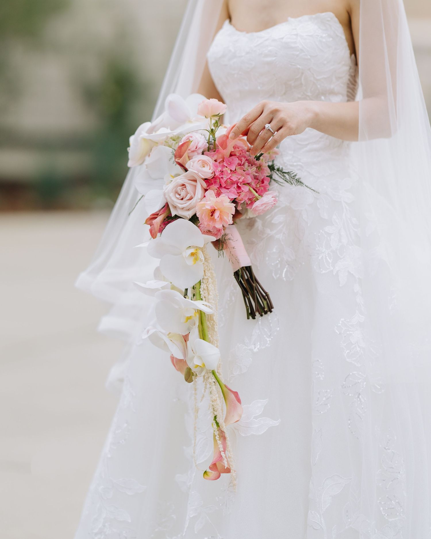 Bride in strapless lace gown holds a cascading bouquet of pink roses, orchids, and hydrangeas.