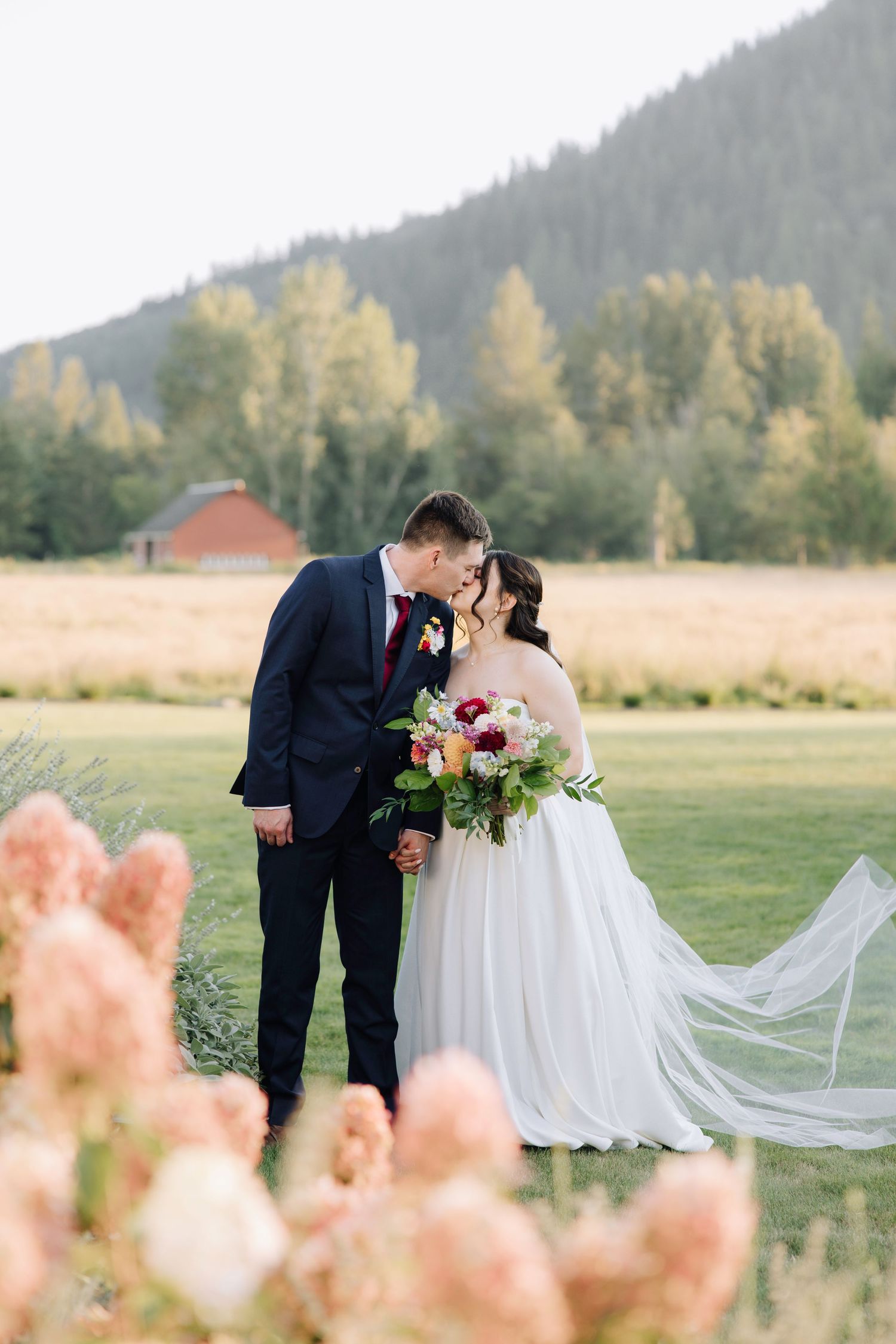 Bride and groom share a romantic moment in a lush green meadow with mountain backdrop and colorful bouquet.