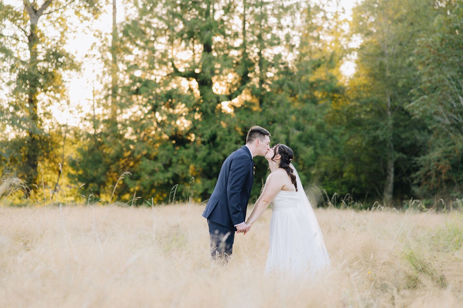 Couple sharing a kiss in a golden field at sunset, bride in white gown and groom in navy suit, lush trees behind.