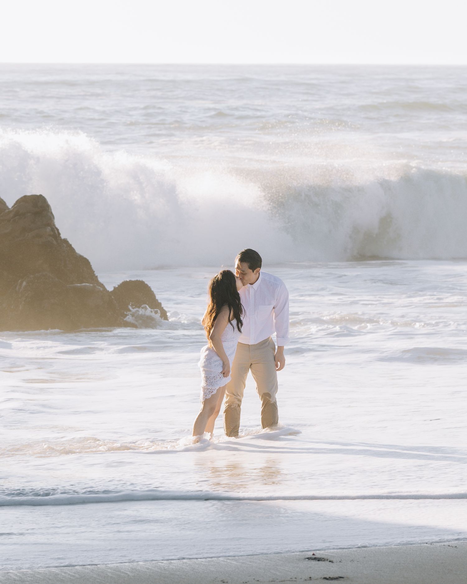 Couple standing close together on wet shoreline with large ocean waves crashing behind at Santa Cruz engagement photoshoot.