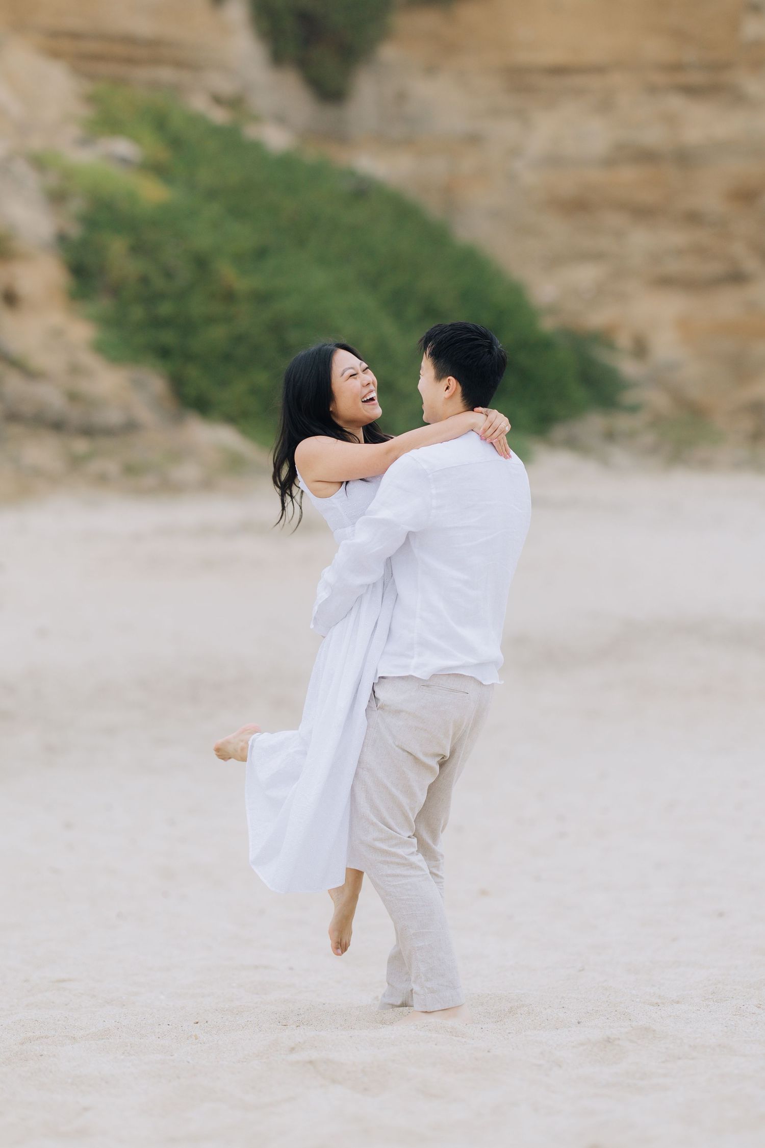 Joyful man lifting smiling woman in white dress on a sandy beach with rocky cliffs during Half Moon Bay beach engagement photoshoot.
