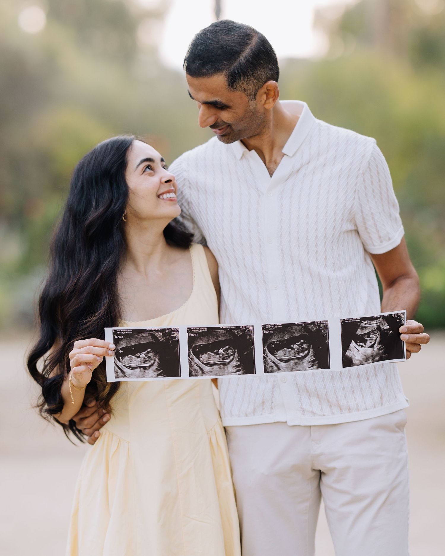 Expecting couple smiling at each other while holding ultrasound photos outdoors at Stanford Arizona Garden maternity photoshoot.