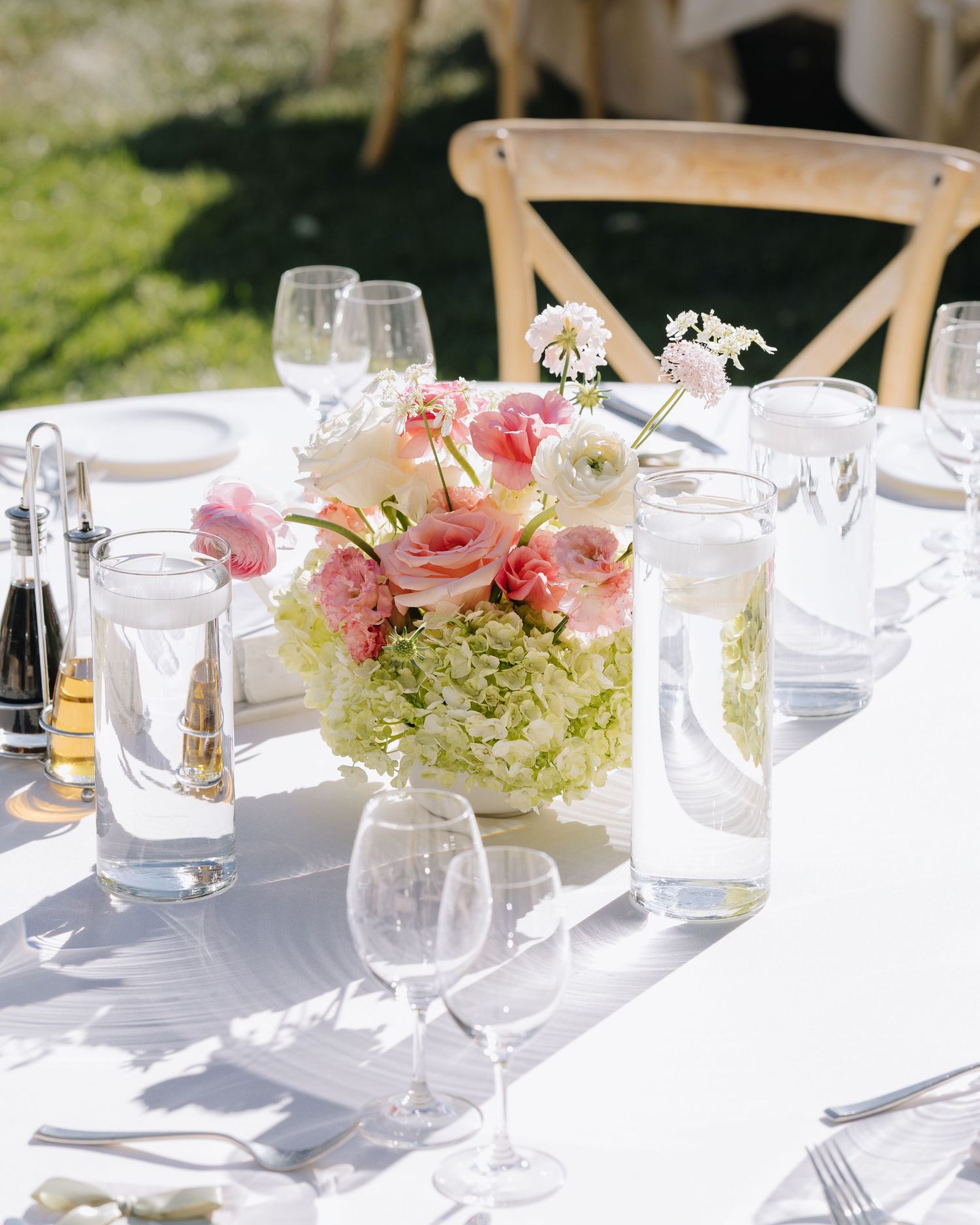 Low centerpiece of pink ranunculus and green hydrangeas on white linen reception table in sunlight.