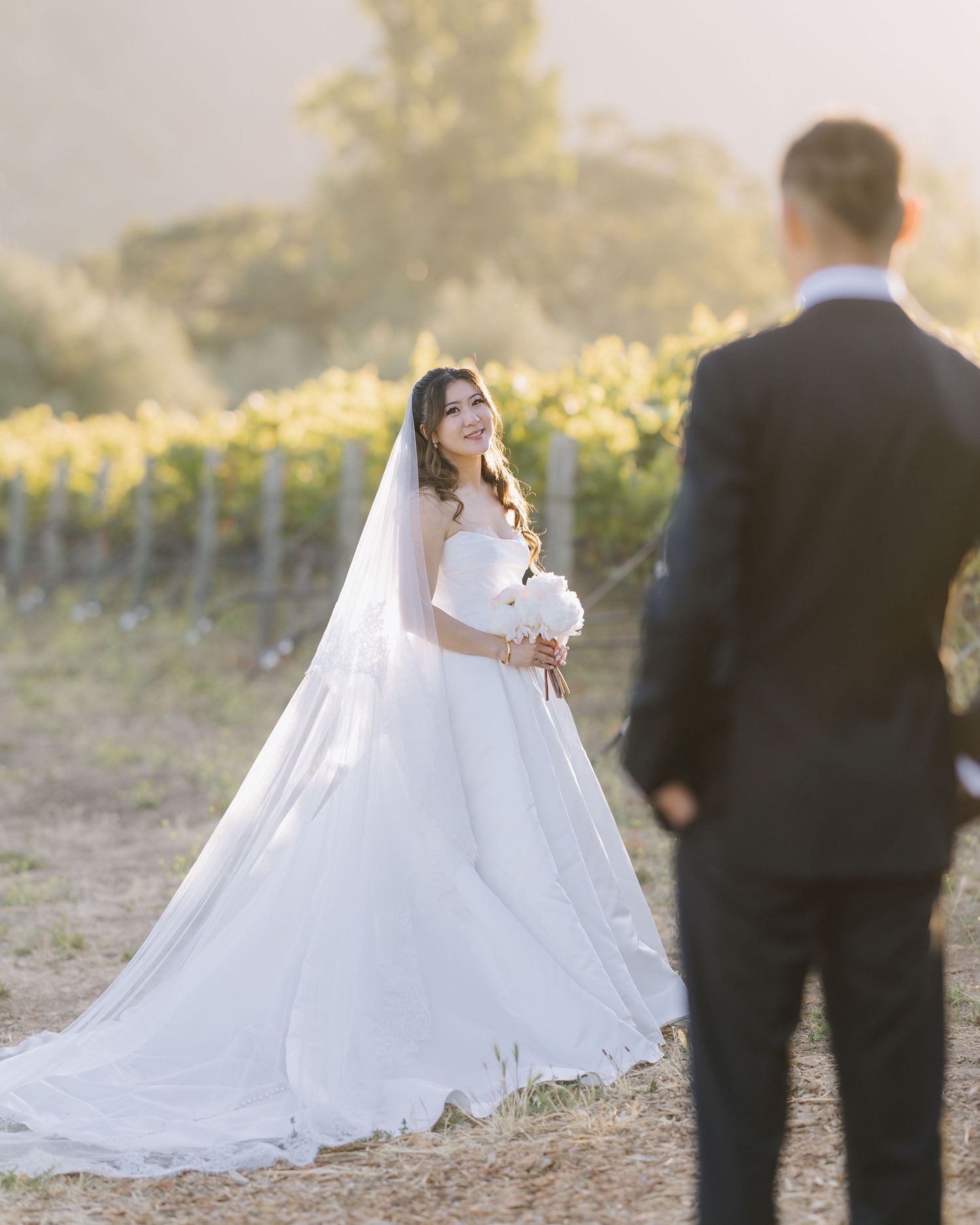 Smiling bride in white ball gown with cathedral veil faces groom at golden hour vineyard at Bernardus Lodge in Carmel Valley, CA.