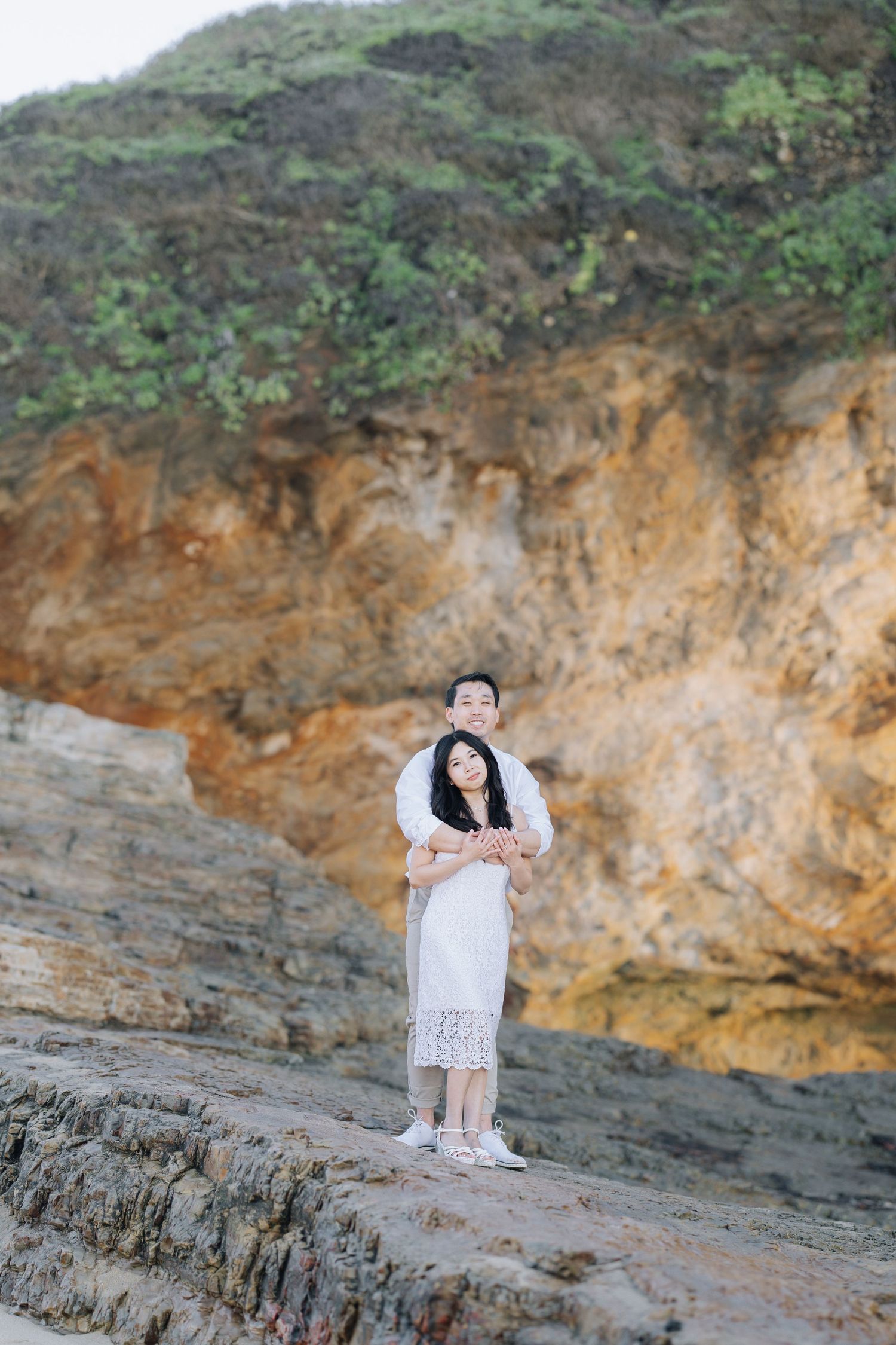 Couple embracing on rocky coastal cliffside with lush green hillside in background at Santa Cruz engagement photoshoot.