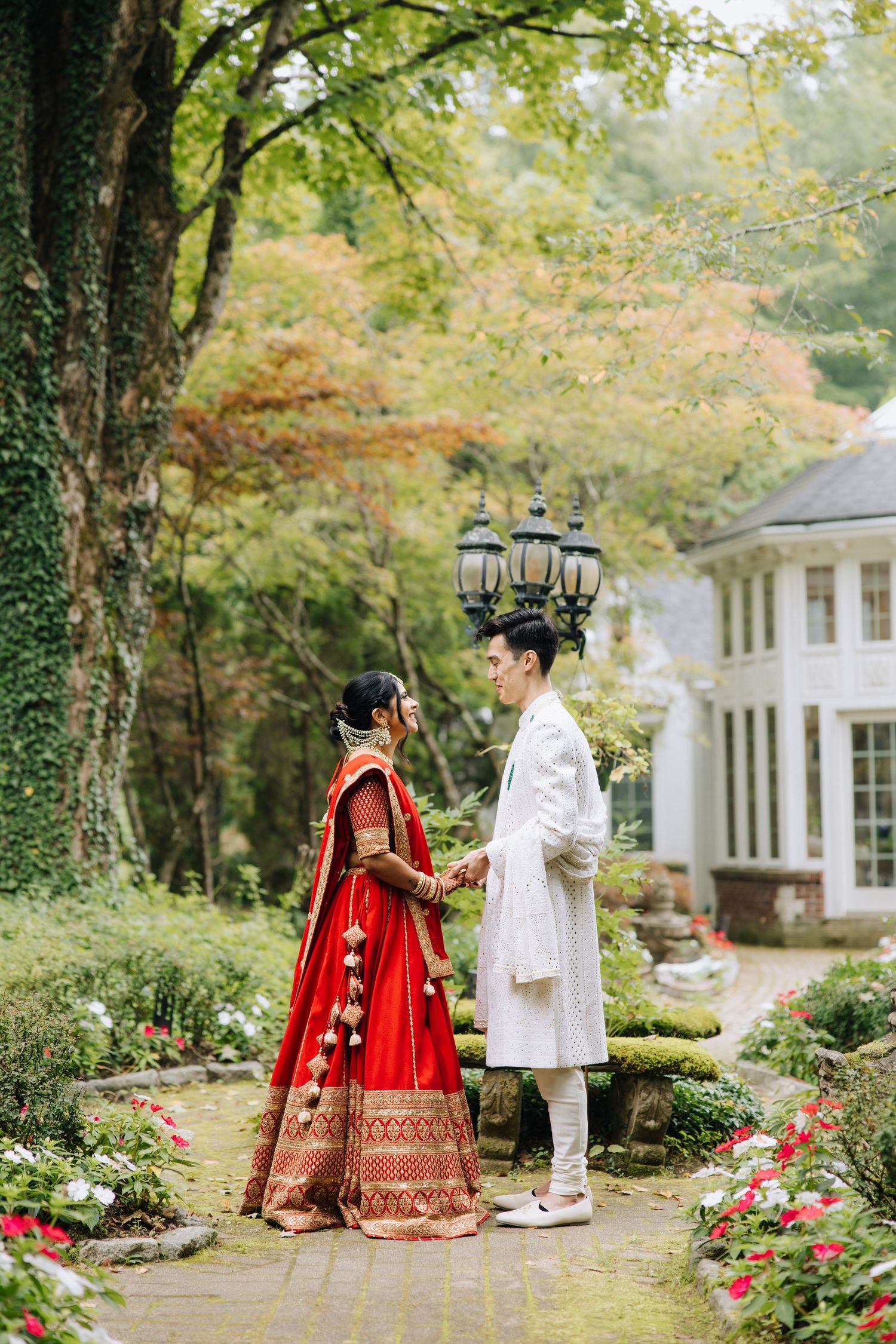 Indian bride in red lehenga and groom in white sherwani hold hands in a lush garden setting.