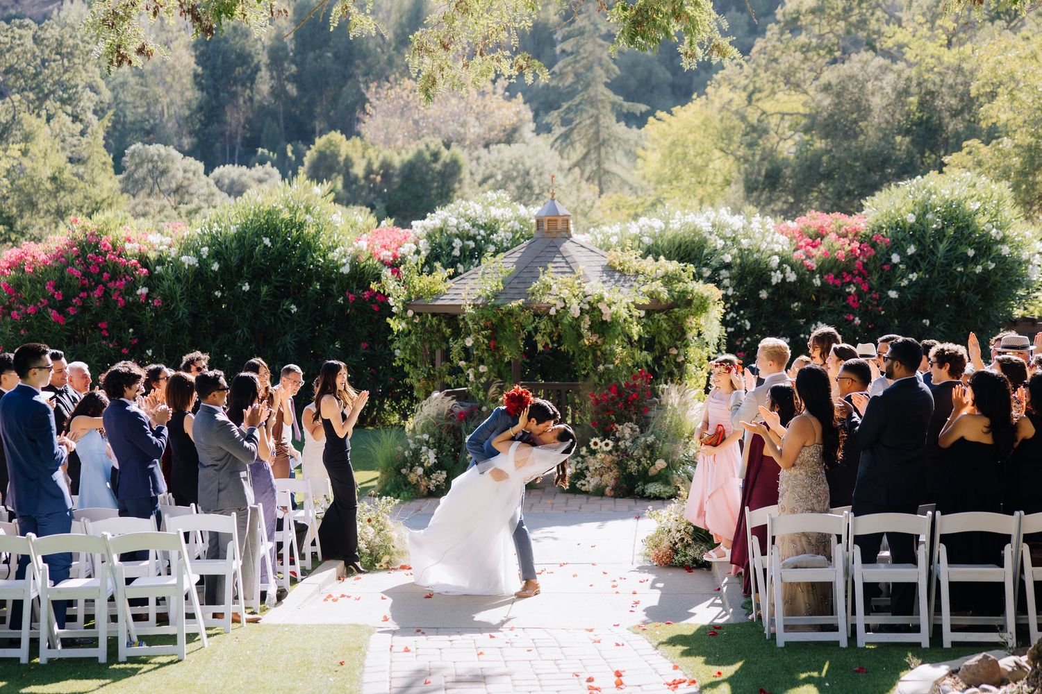 Bride and groom share first kiss at outdoor wedding ceremony surrounded by guests and floral arch at Elliston Vineyards in Sunol, California