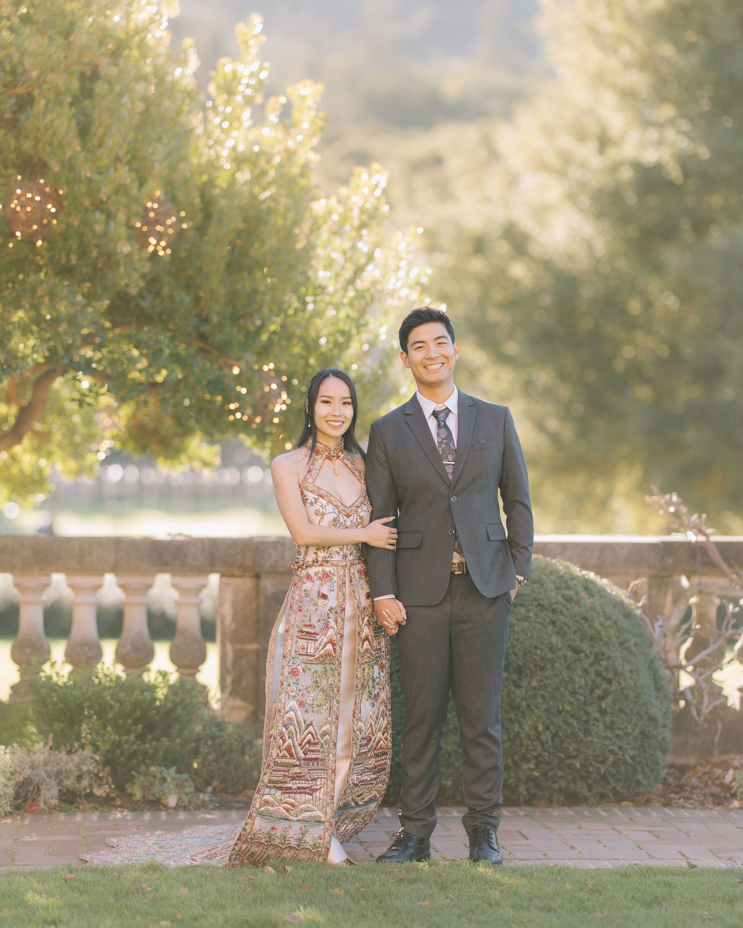 Smiling couple in elegant formal wear standing together in golden-hour garden with string lights during Filoli engagement photoshoot.